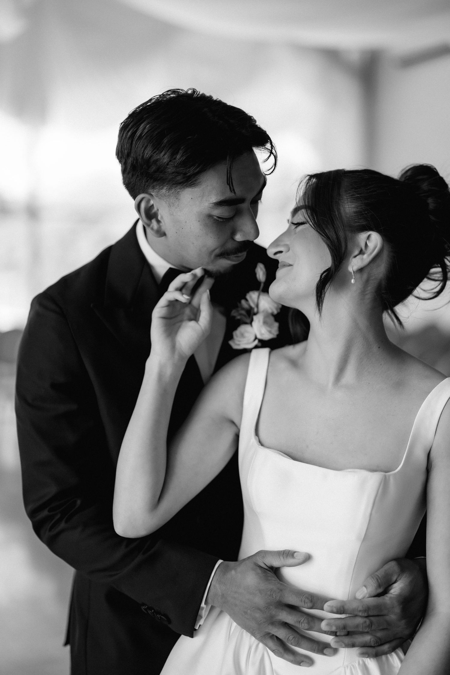 In a black and white portrait, a bride in a square-neck gown and a groom in a tuxedo share an intimate, affectionate moment.
