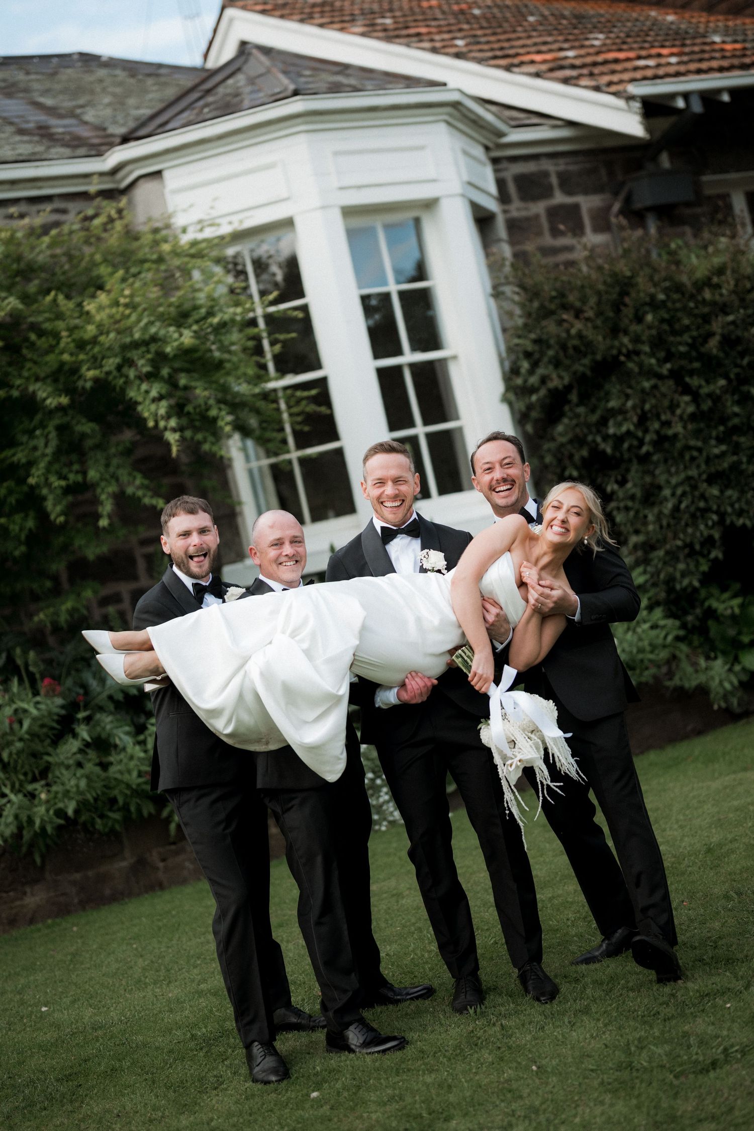 Groomsmen in black tuxedos playfully carrying someone in a white dress across the lawn.