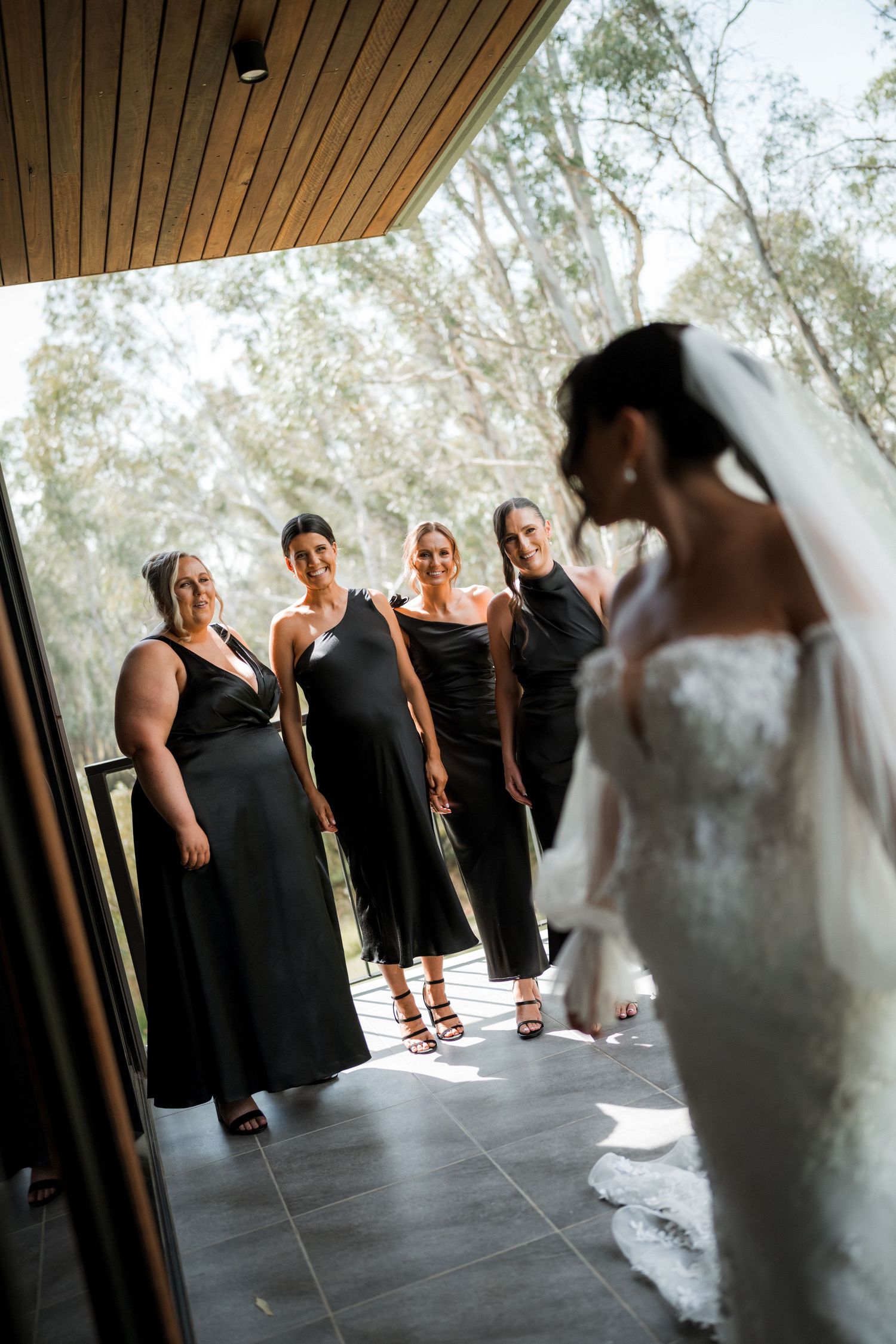 A wedding party in black dresses stands in a row while a bride in white approaches them from behind.
