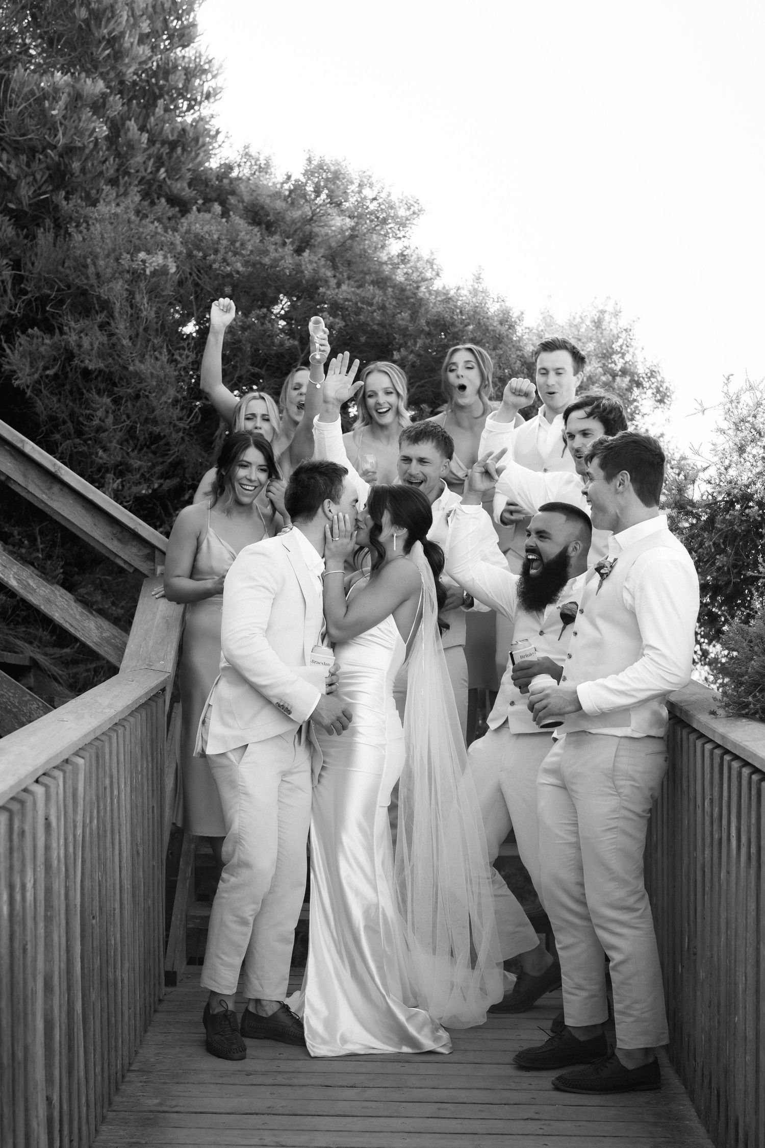 Wedding party celebrates with playful poses and laughter on a wooden boardwalk in sequence of black and white photos.