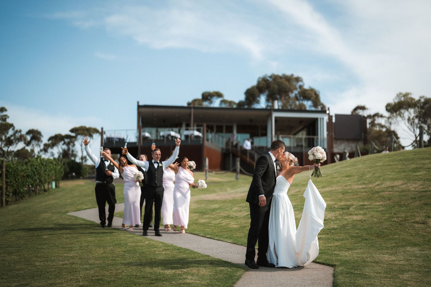 Wedding party gathered on a hilltop path with modern building and blue sky in background.