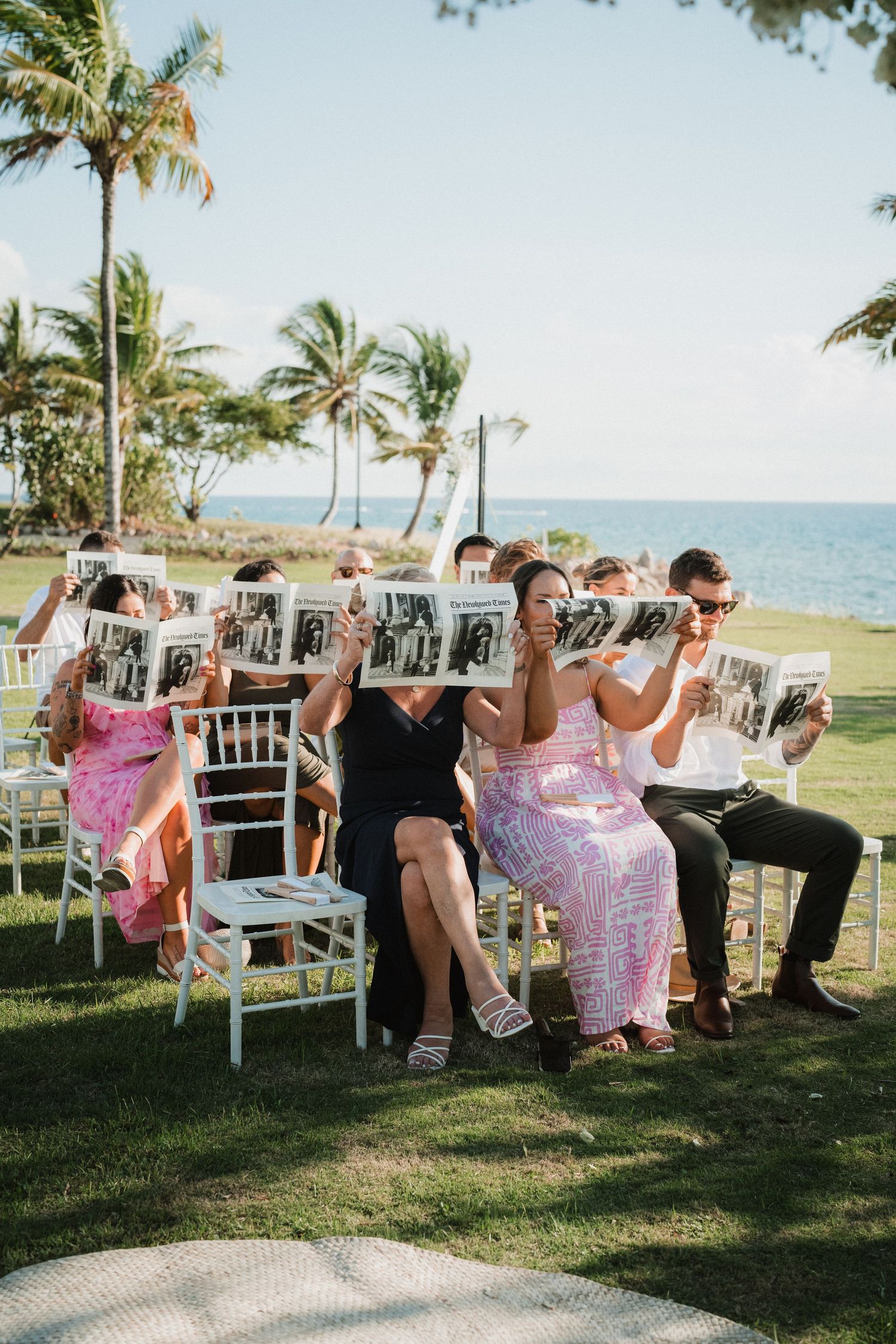 Wedding guests hold up personalized fans during an outdoor beach ceremony surrounded by palm trees.