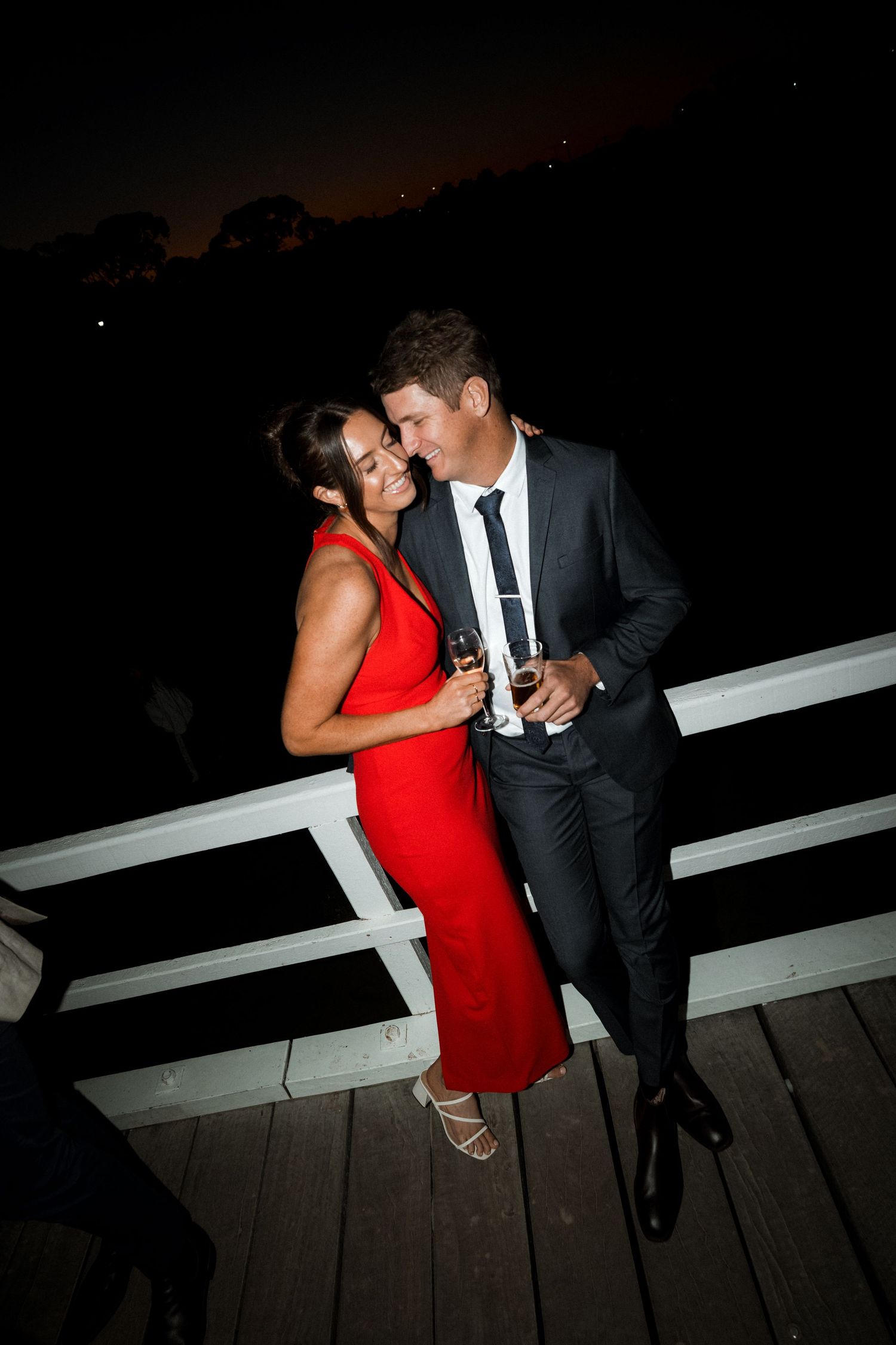 A couple sharing an intimate moment on a dark deck, with a woman in a red dress and man in a suit.
