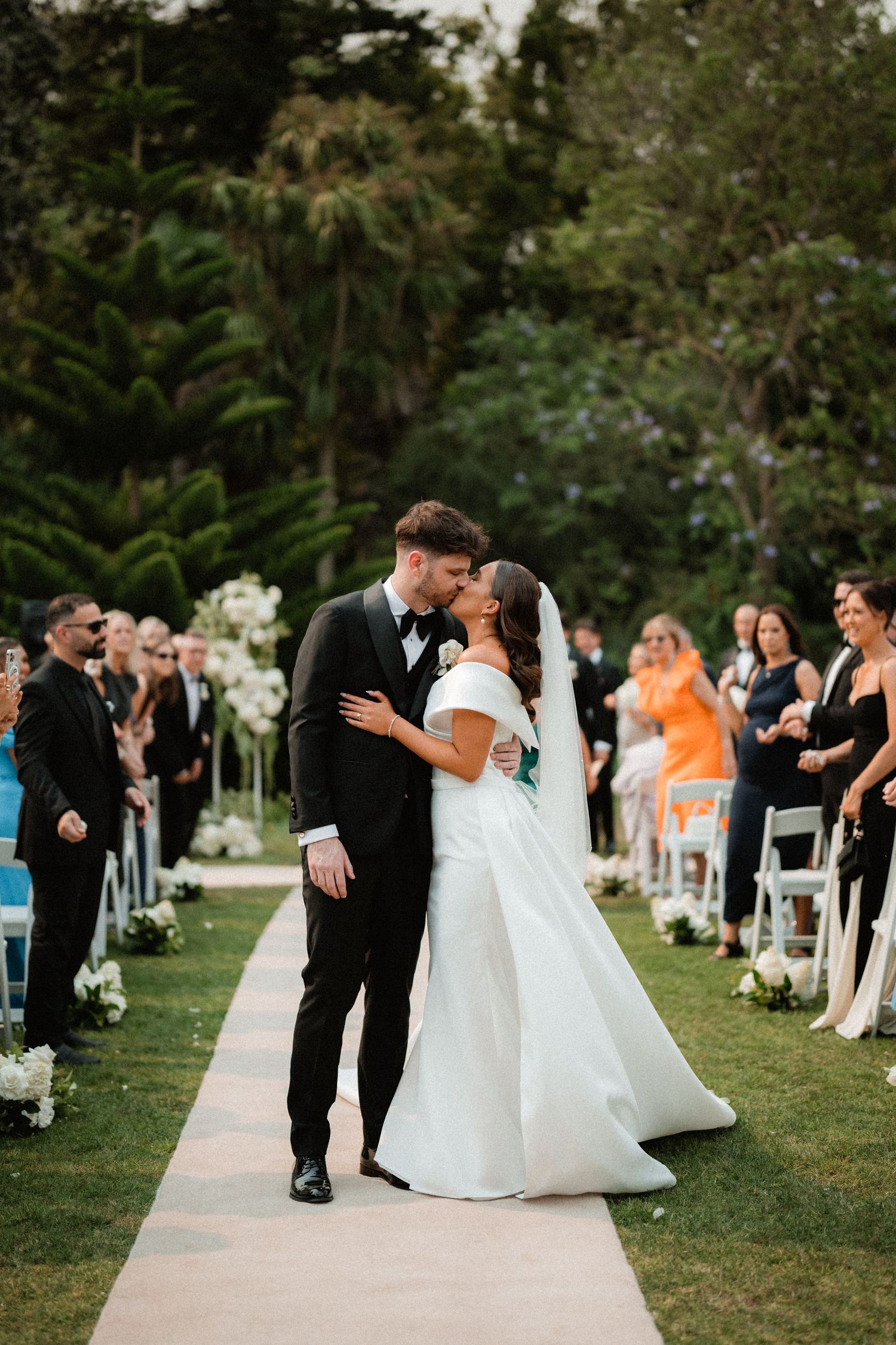 A couple sharing their first kiss as newlyweds on a white pathway surrounded by wedding guests.