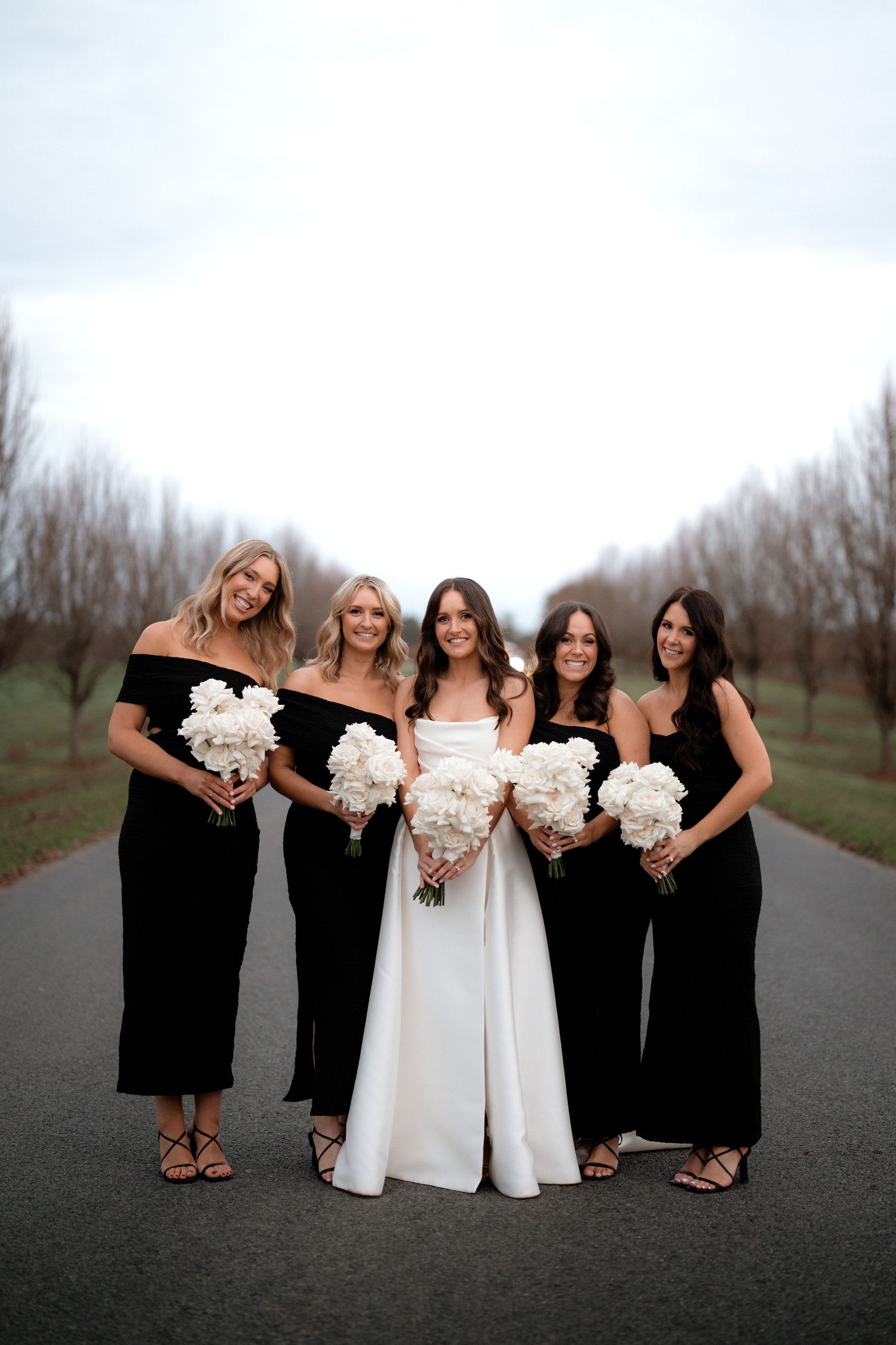 Wedding party poses on path with bride in white gown and bridesmaids in elegant black dresses.