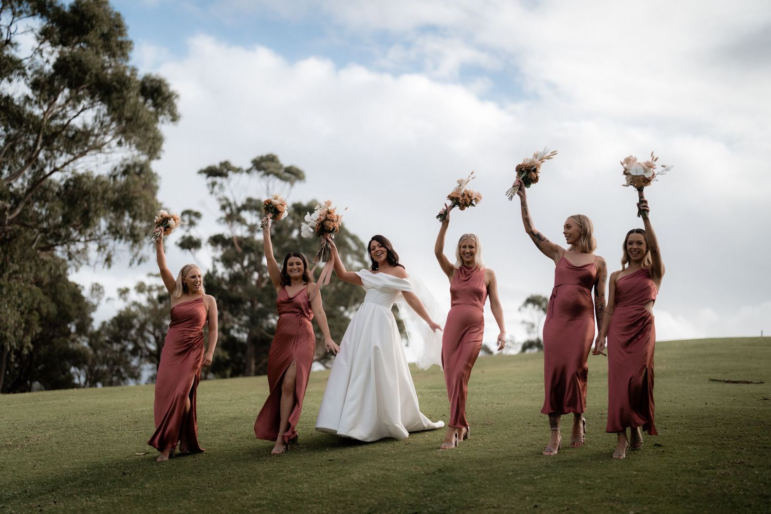 A bridal party in dusty rose dresses joyfully celebrates on a grassy hill beneath cloudy skies.