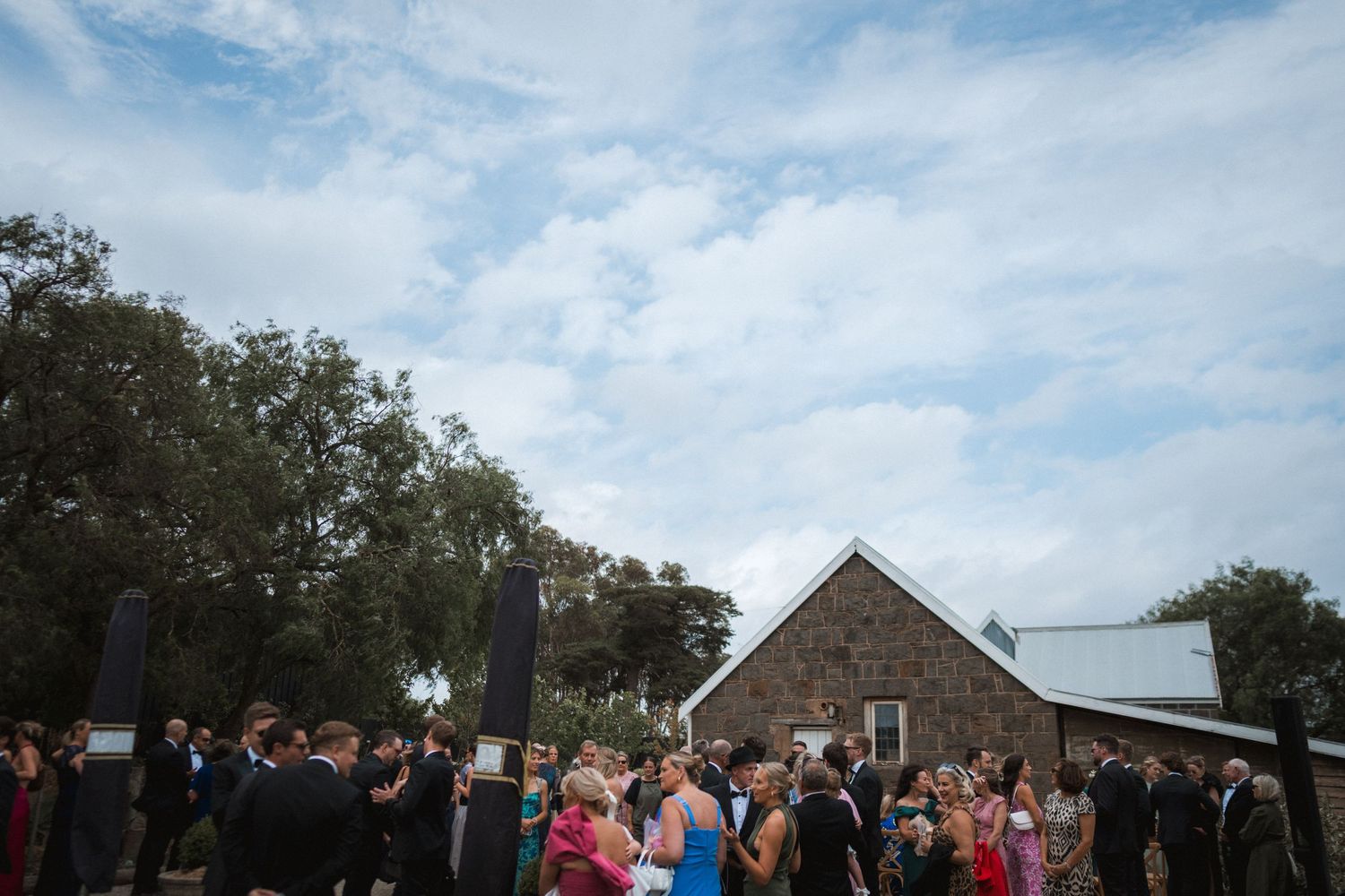 Wedding guests gather outside a rustic brick church building under blue skies and oak trees for an outdoor ceremony.
