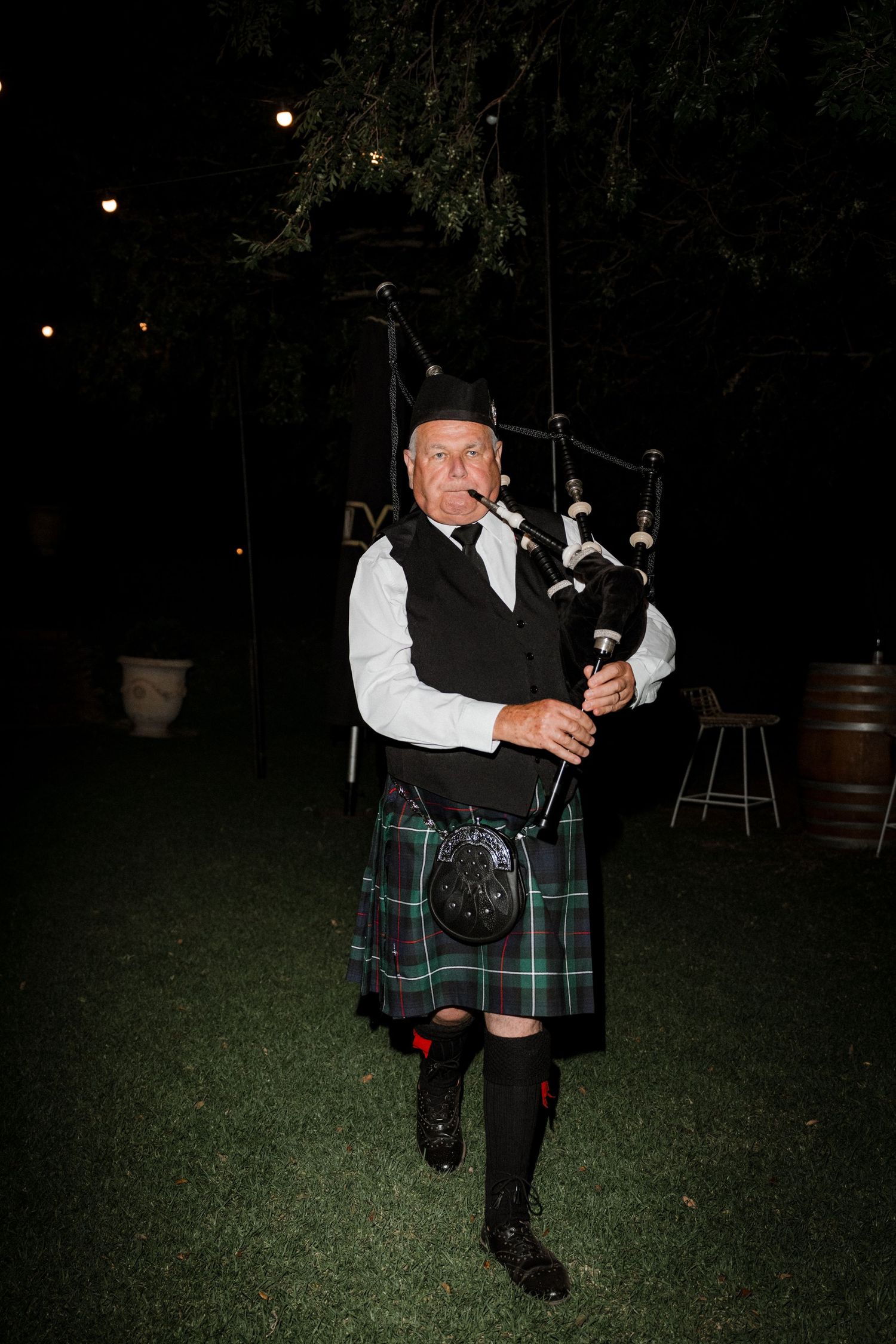 Traditional bagpiper in formal Scottish attire performs outdoors at night on grass with barrel decor in background.