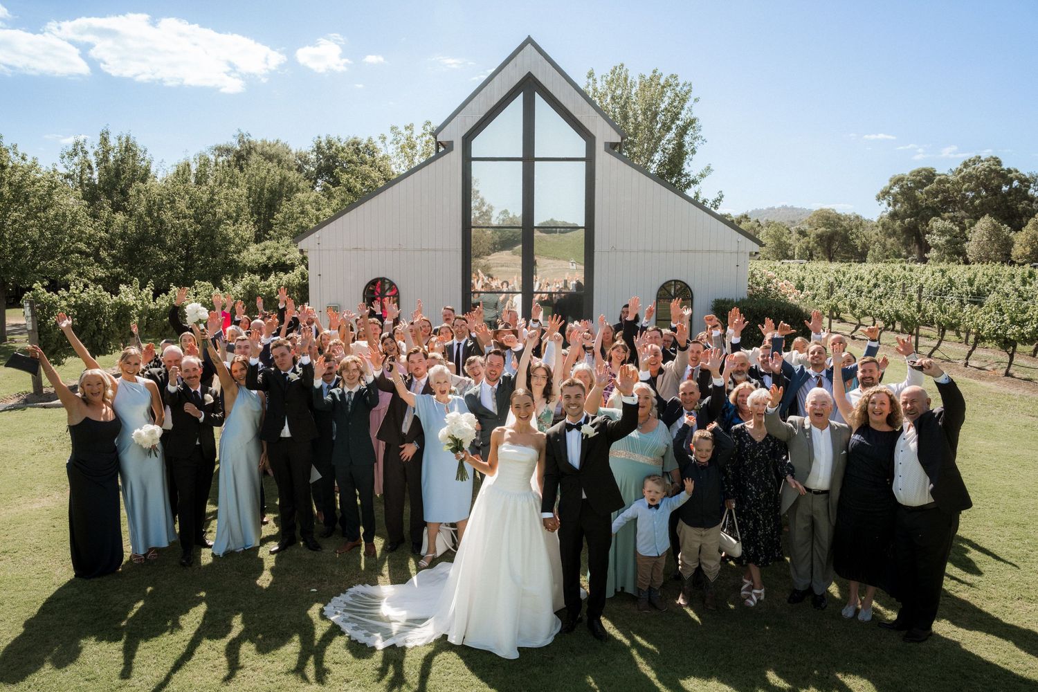 Large wedding party poses for group photo outside modern chapel with glass windows on sunny day.