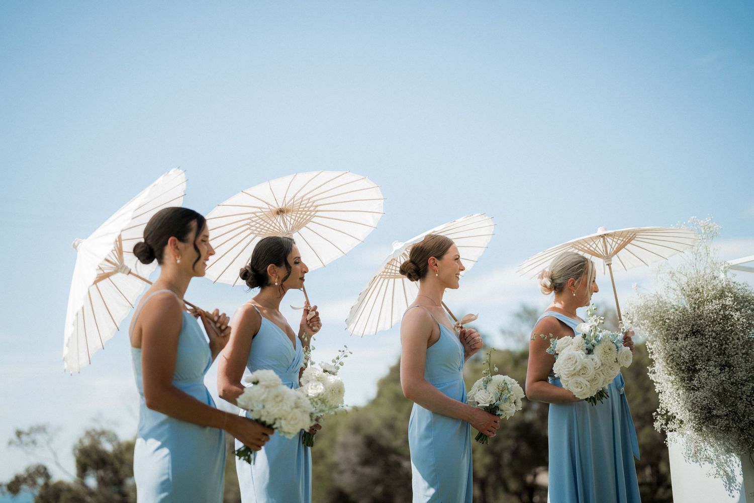 Bridesmaids in light blue dresses holding white parasols against a bright blue sky.