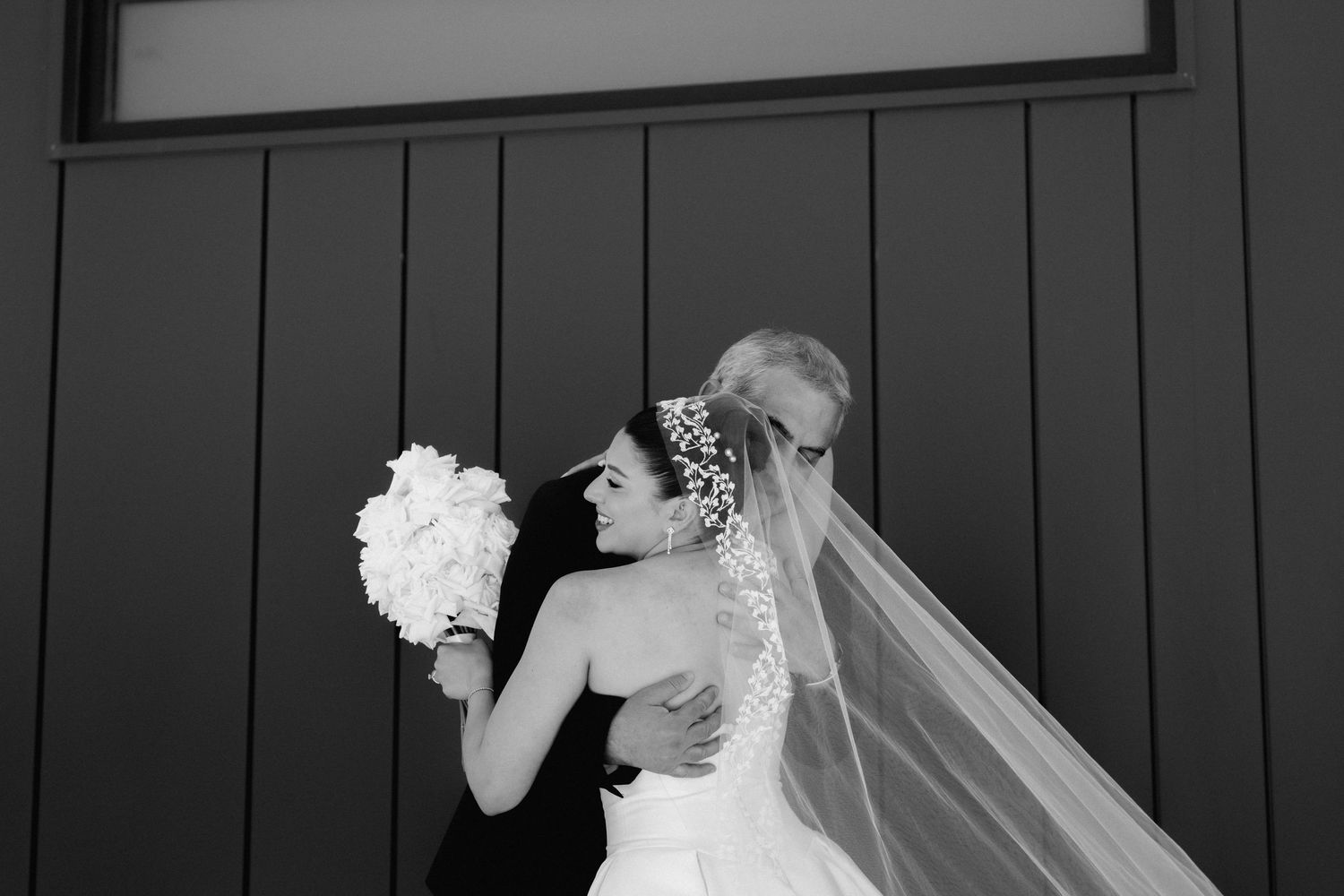 A bride in a white wedding dress and veil stands against a dark paneled wall in a dramatic black and white photo.