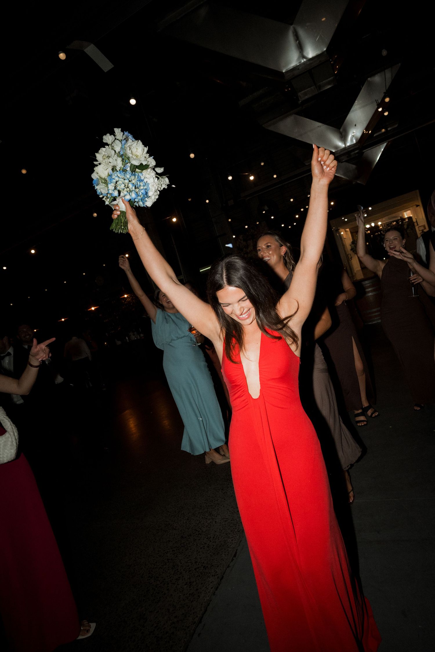 Woman in red dress celebrates joyfully with raised arms holding a blue and white bouquet.
