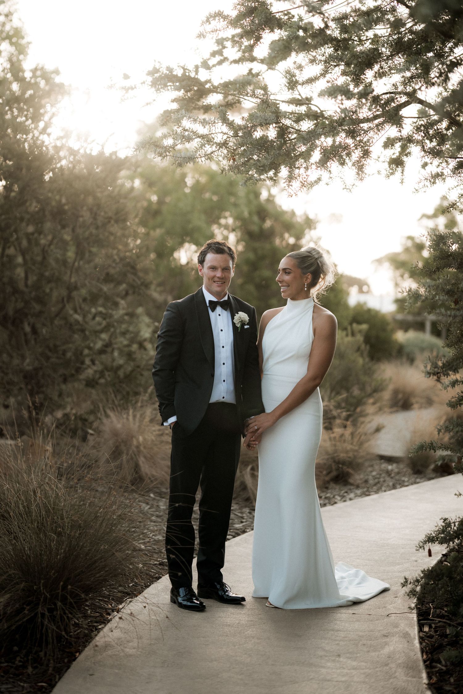 Wedding couple posing on outdoor path surrounded by trees at sunset.
