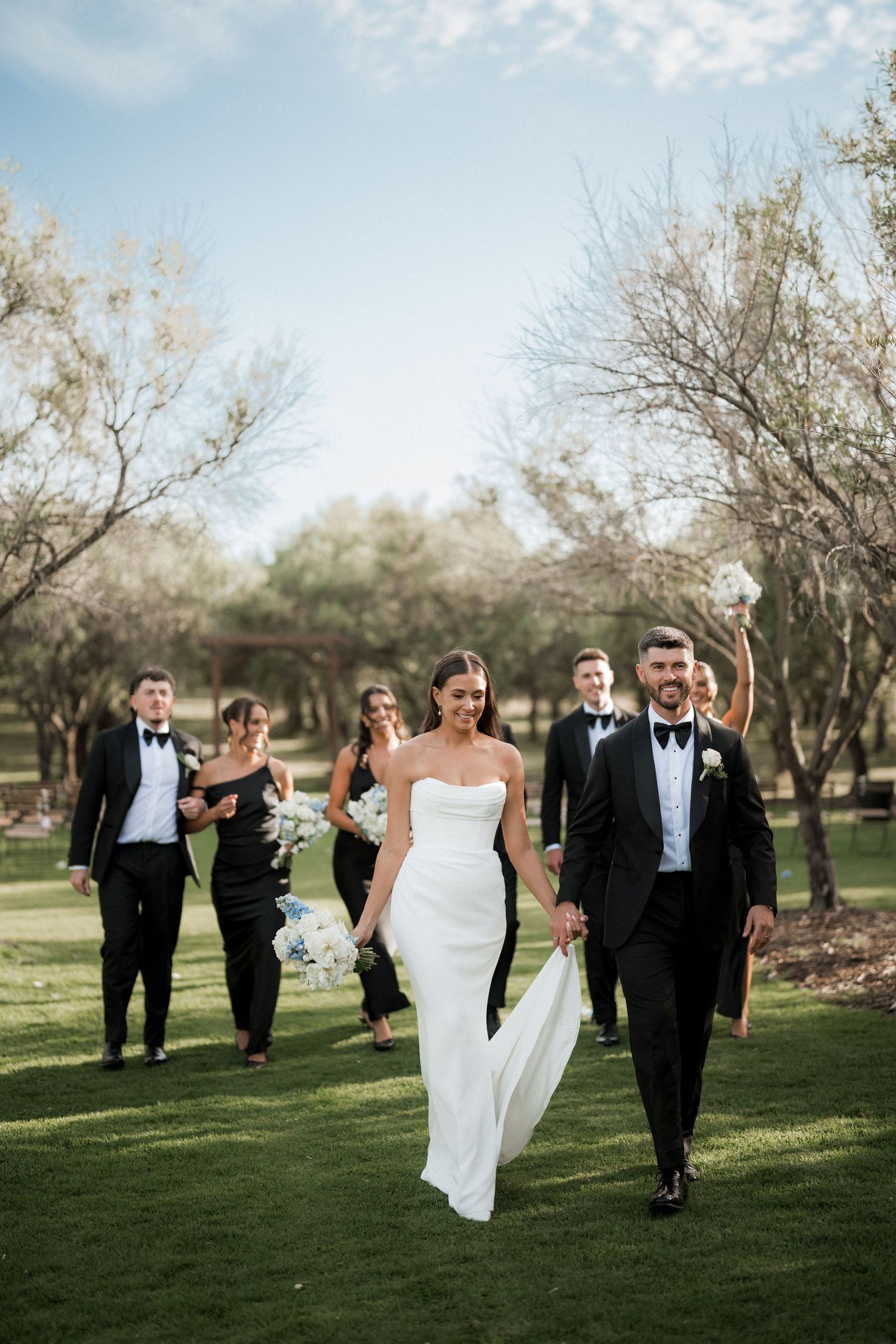Wedding party walking and celebrating on a grassy lawn with trees in the background.