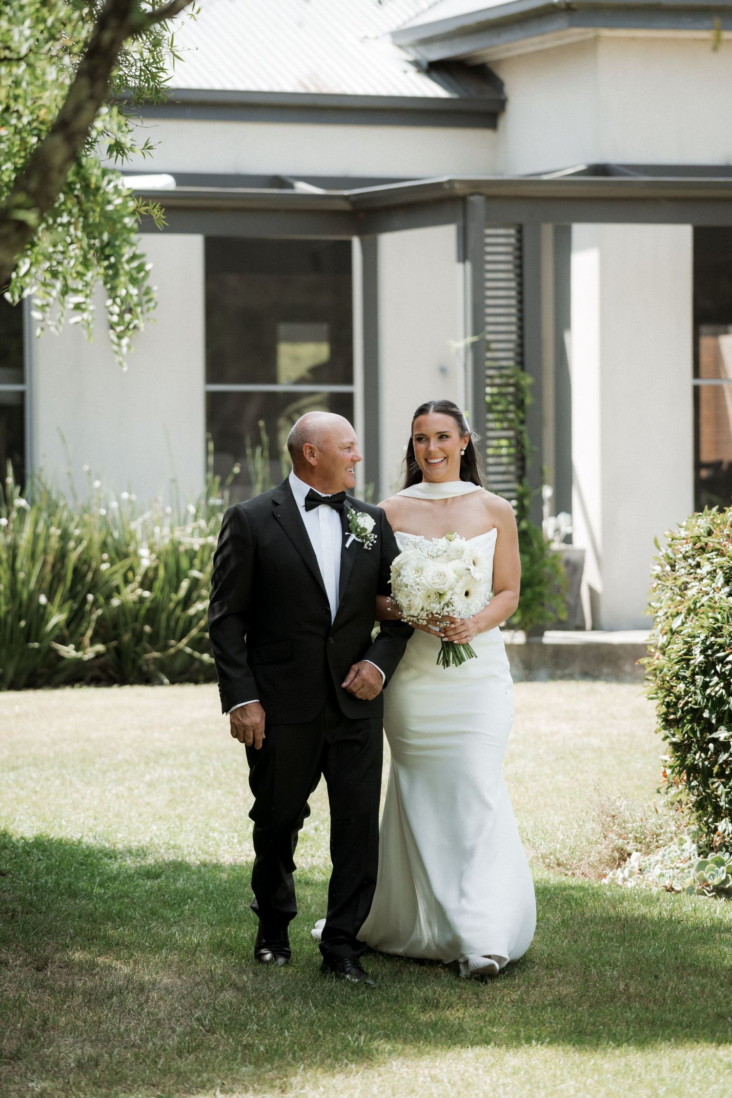 A wedding couple walks together on a sunny lawn outside a modern white building.