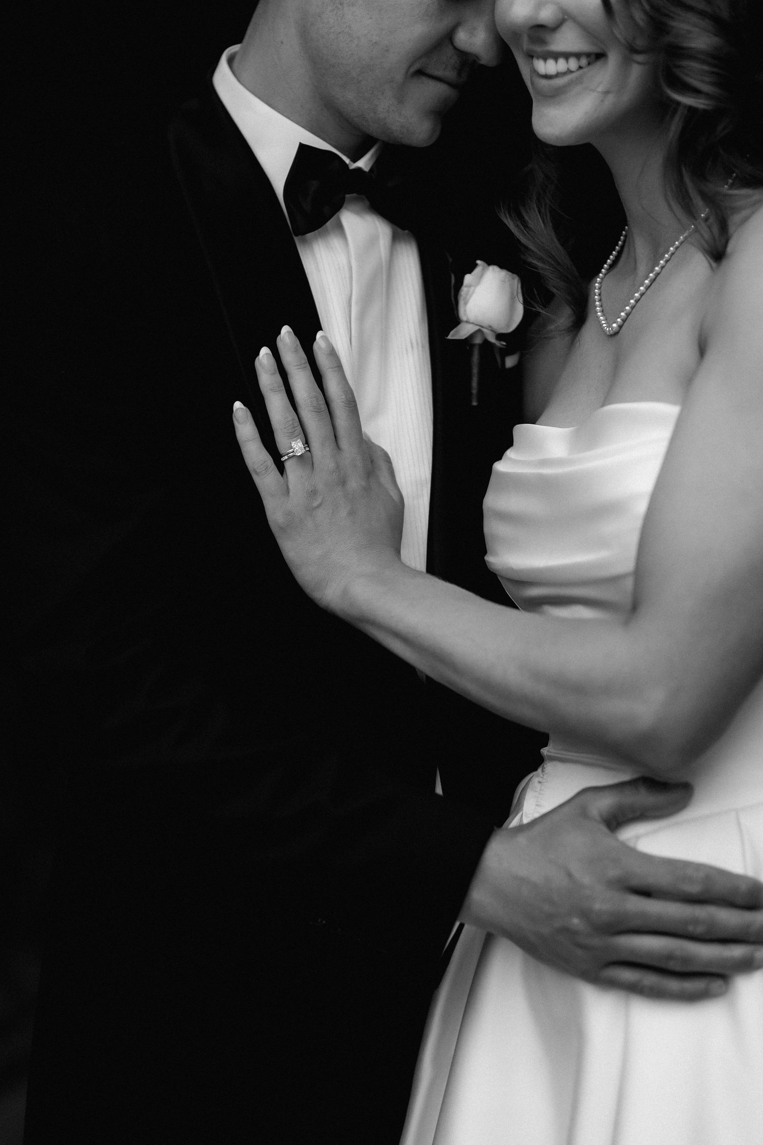 A black and white close-up wedding photo showing a couple embracing, with the bride in white and groom in tuxedo with bowtie.