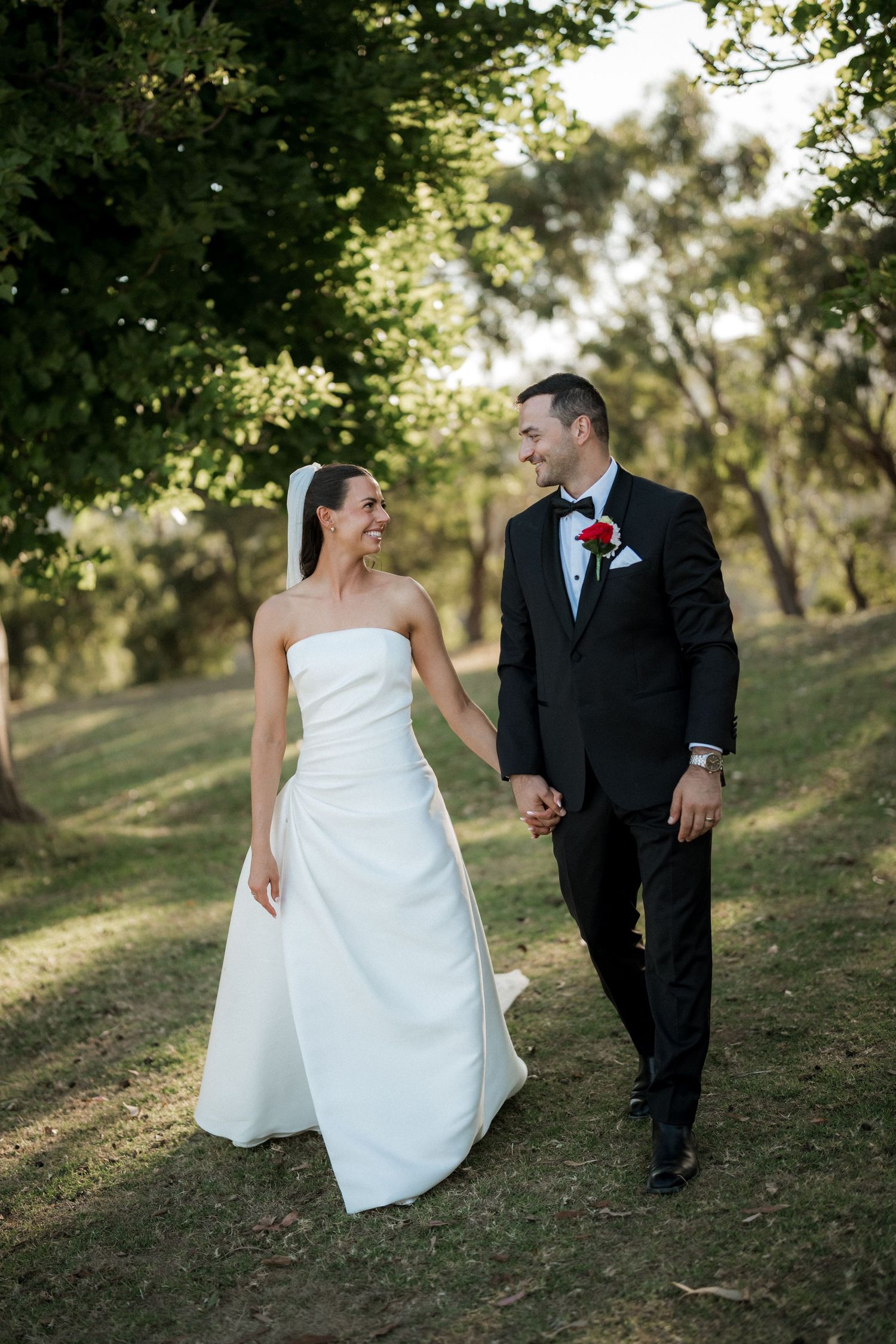 Newlyweds walk hand in hand along tree-lined path during outdoor wedding photoshoot.
