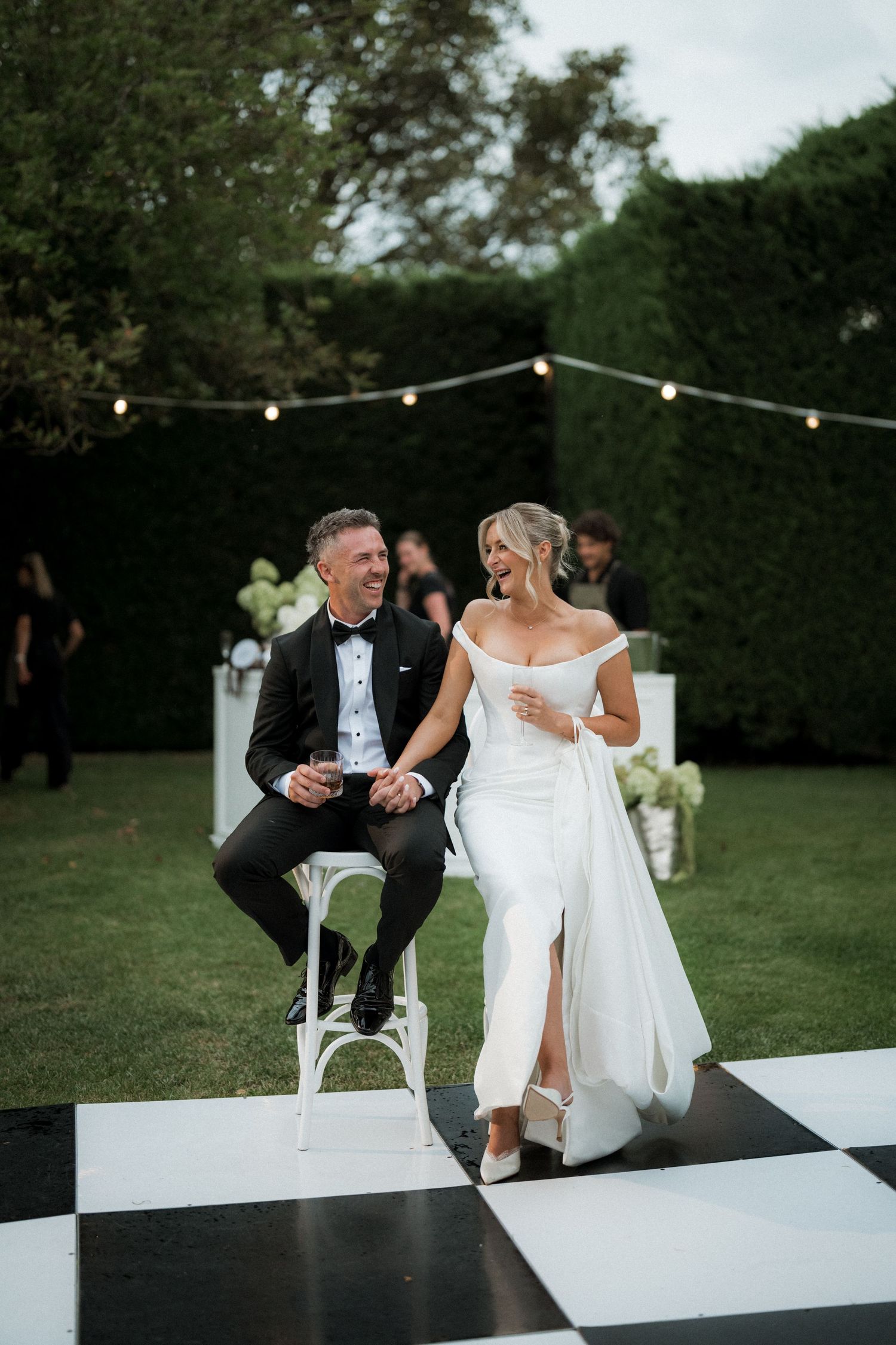 Wedding couple sits on modern white platform during outdoor evening reception.