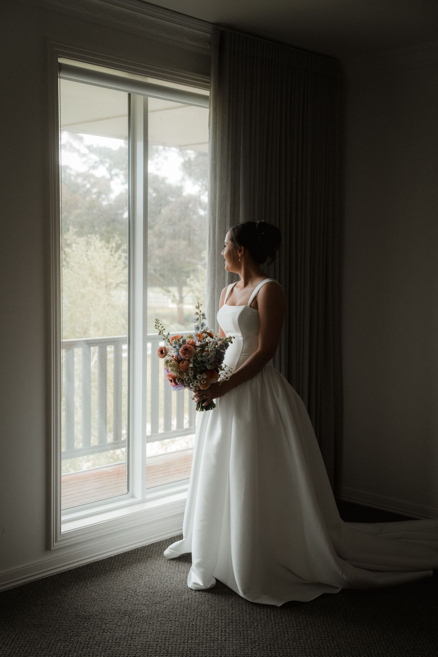 Silhouette of a bride in flowing white gown looking out large windows in a dim room.