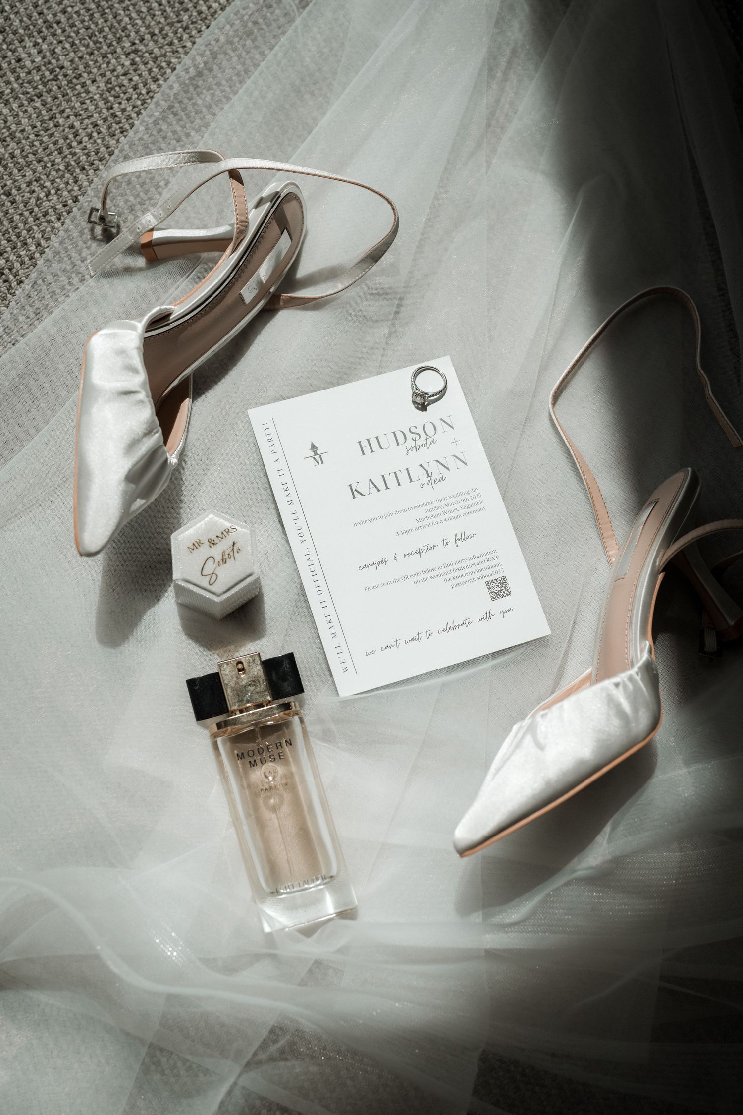 Wedding accessories including white heels, perfume bottle, and invitation card arranged on textured fabric.