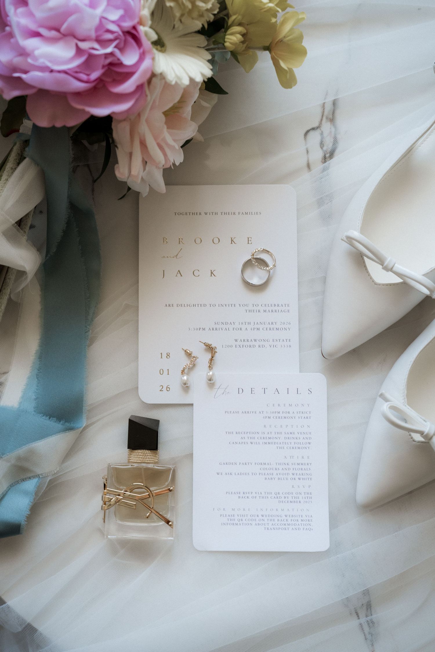 Elegant wedding flat lay with invitation suite, gold rings, pearl earrings, YSL perfume, and white heels on marble.
