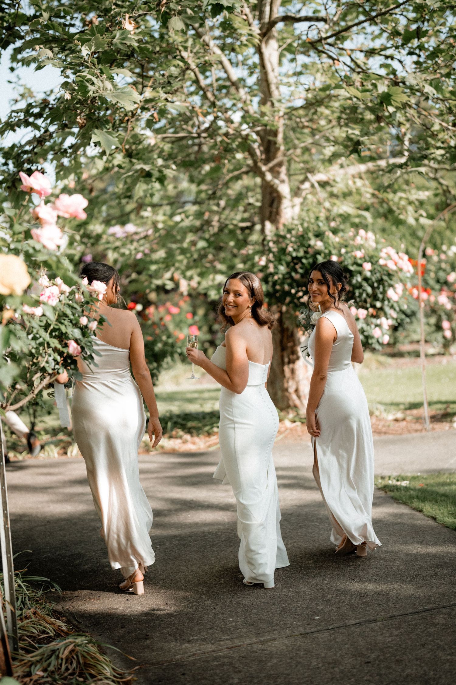 Three bridesmaids in white dresses walking along a garden path surrounded by trees and flowers.