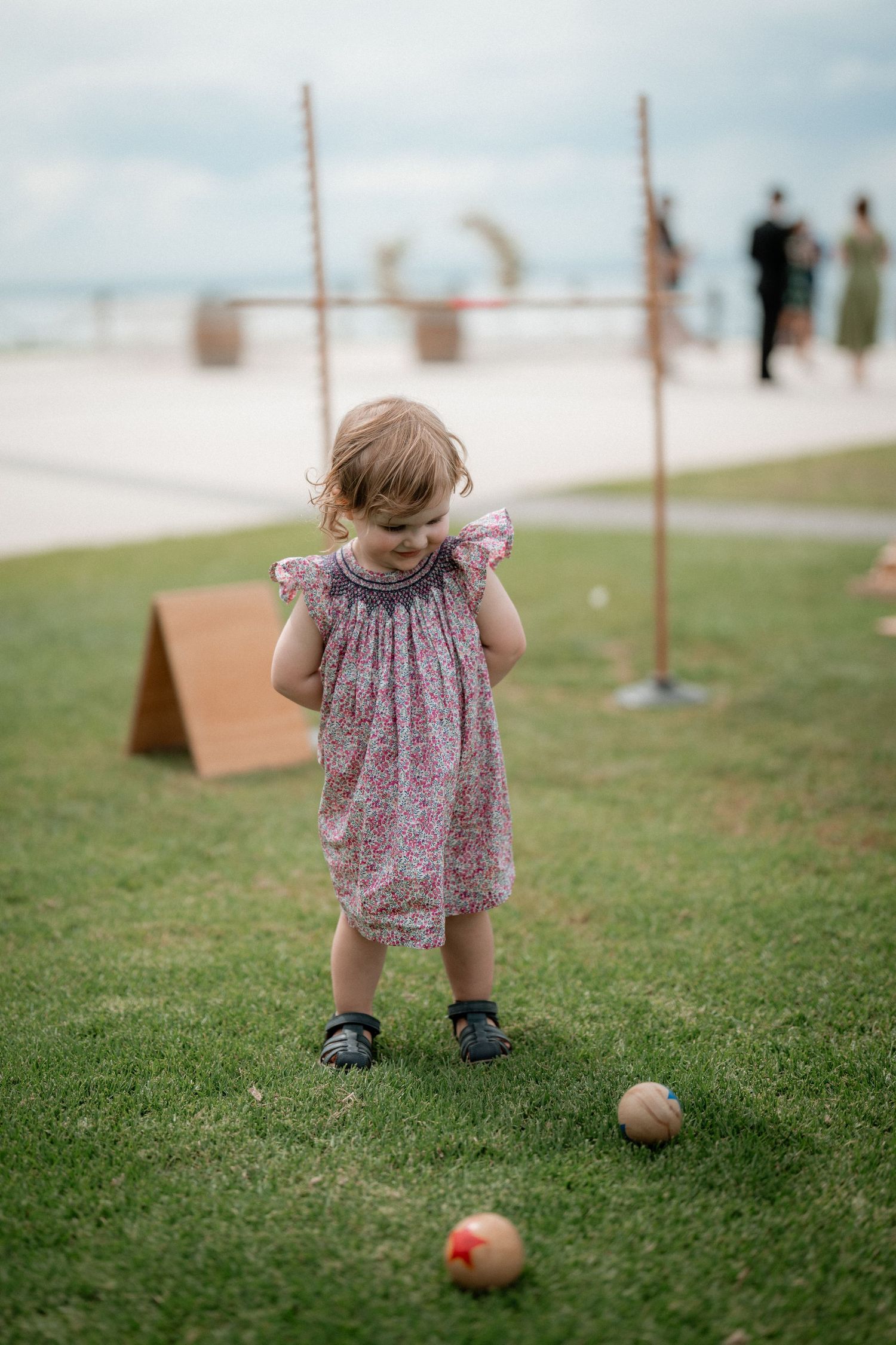 Small child in floral dress and dark shoes plays on grass with colorful balls at an outdoor event venue.