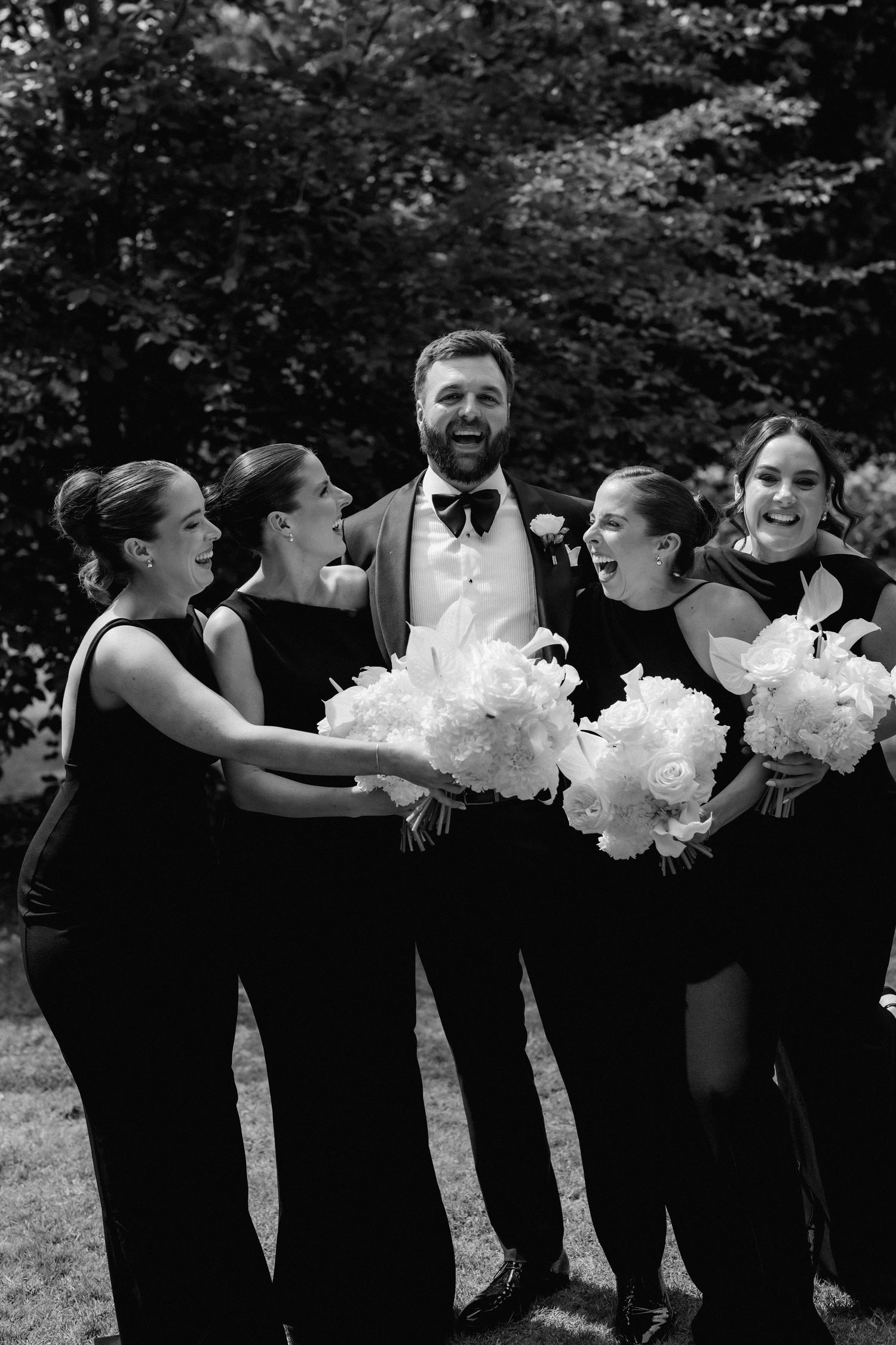 Groom in tuxedo laughs with four bridesmaids in black dresses holding white peony bouquets outdoors.