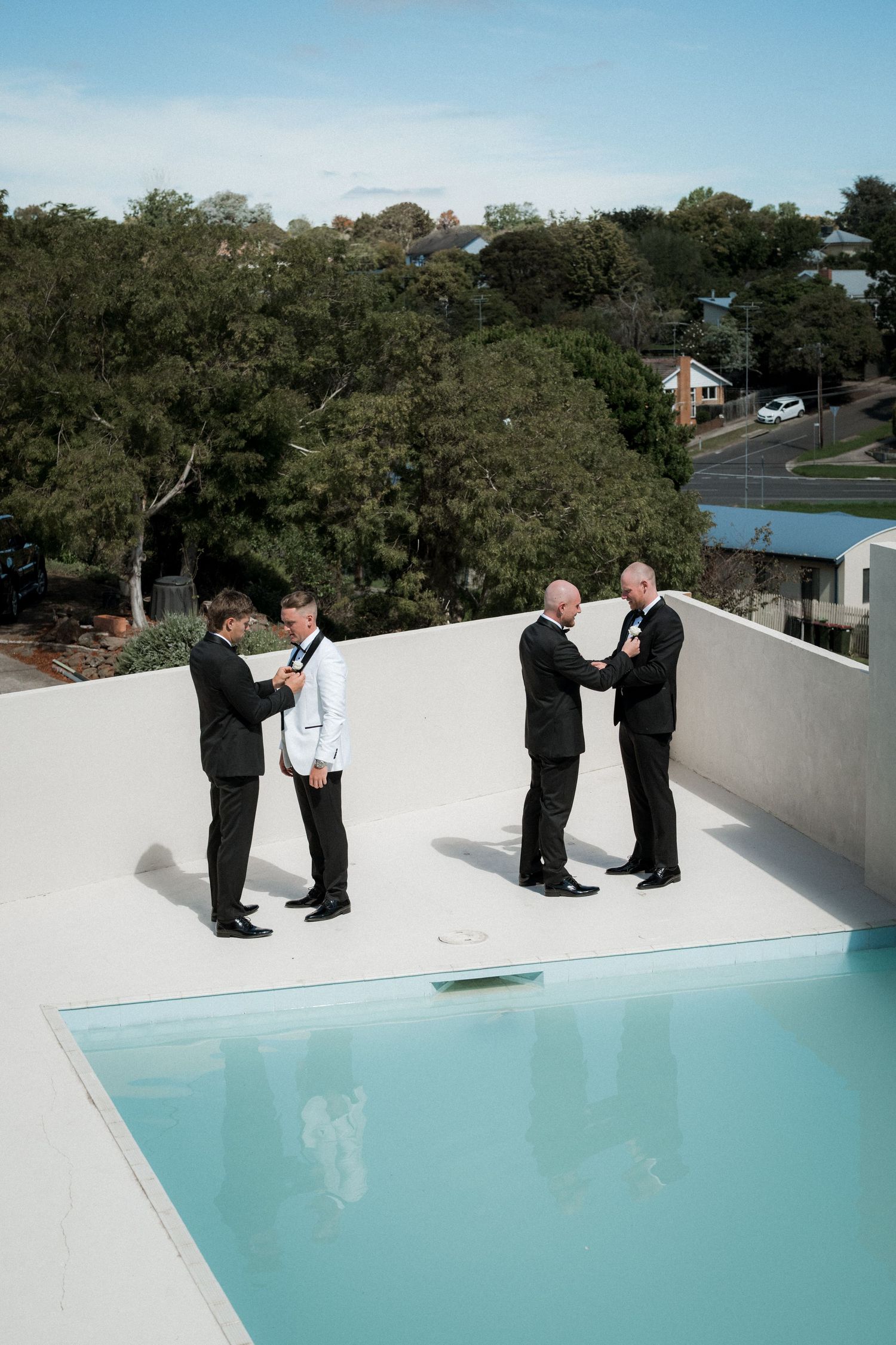 Two people in formal attire shake hands on a white rooftop with a swimming pool and trees in the background.