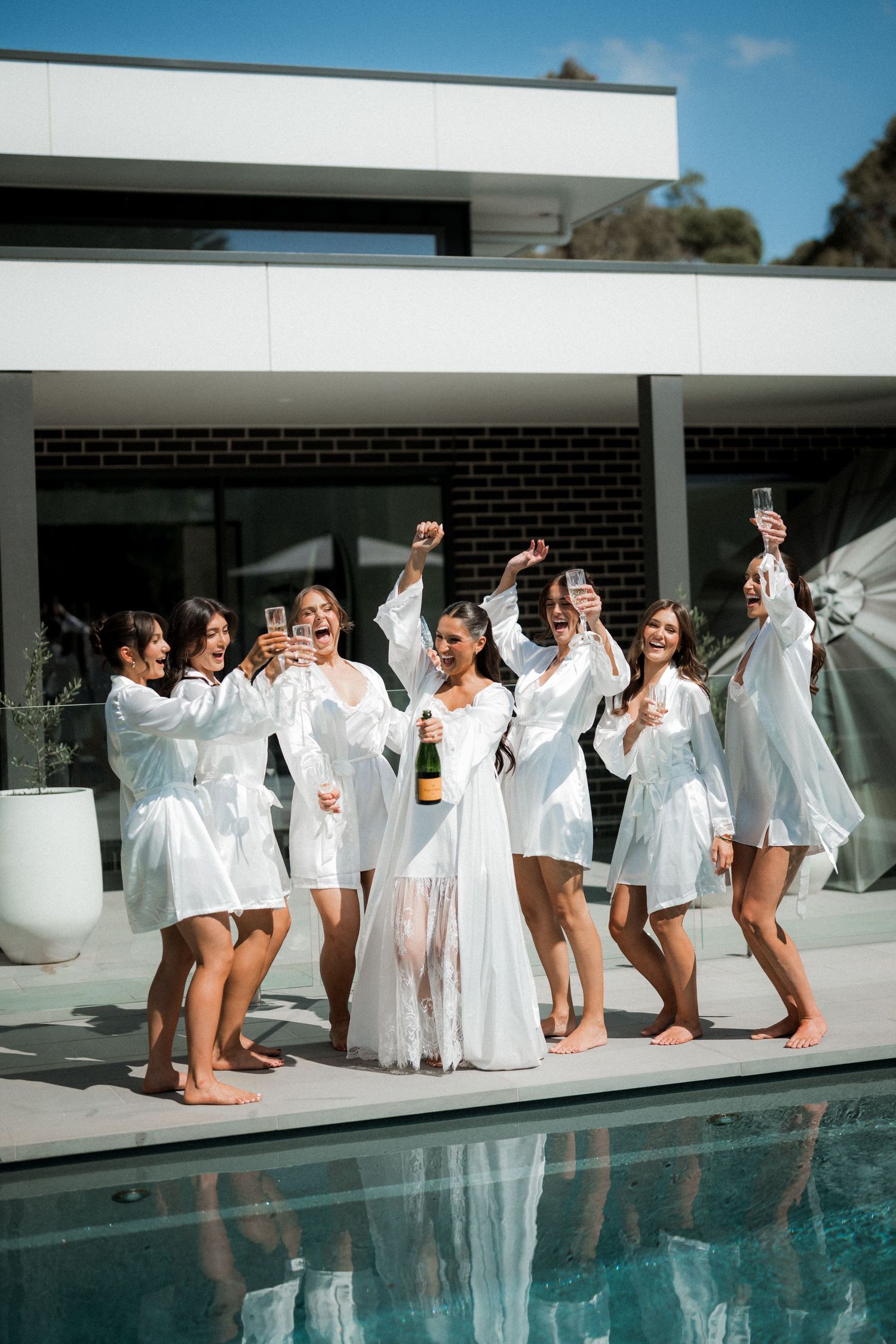 Bridesmaids in white robes pose playfully by a swimming pool on a sunny day.