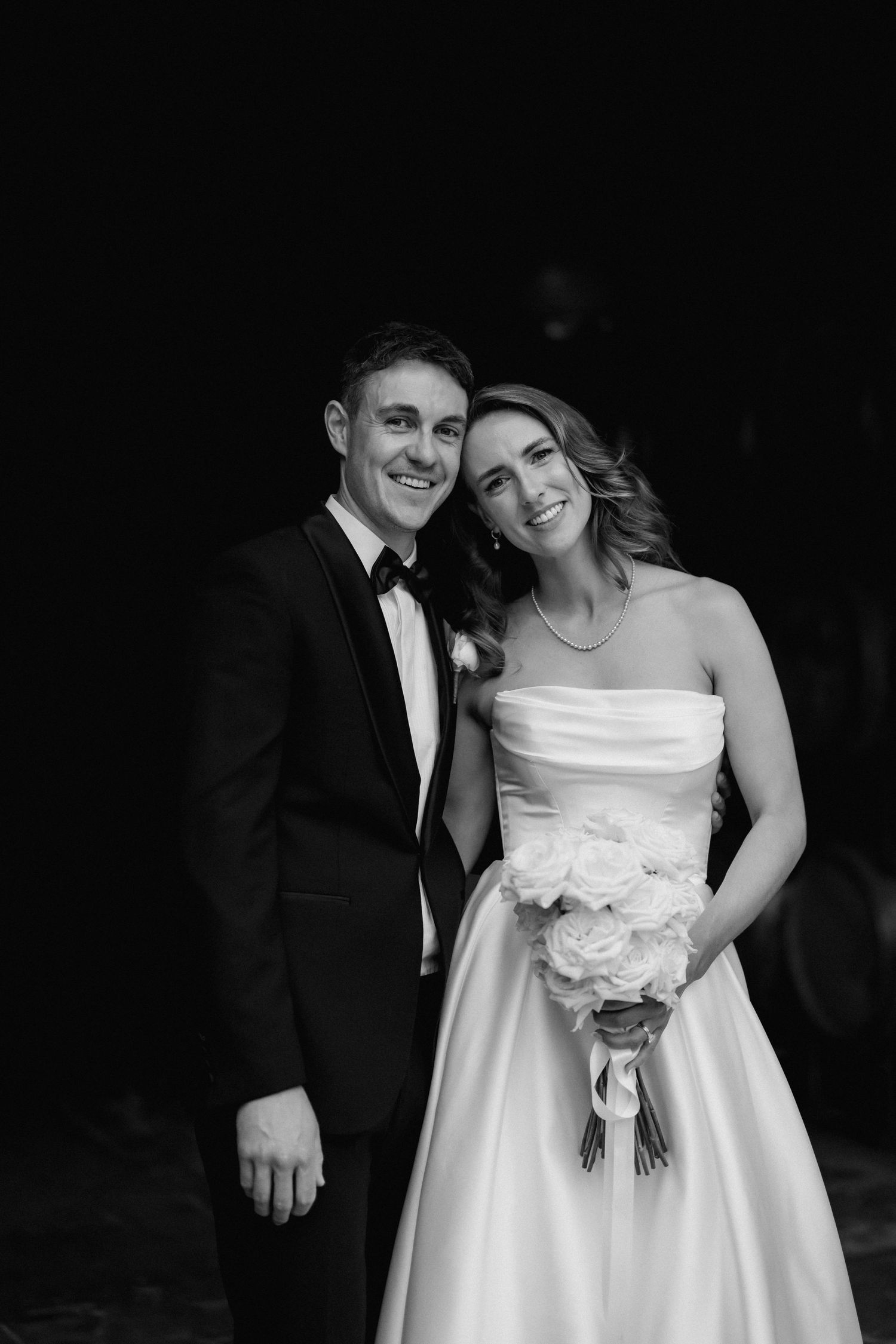 Black and white portrait of newlyweds in formal attire holding a bouquet of white roses.