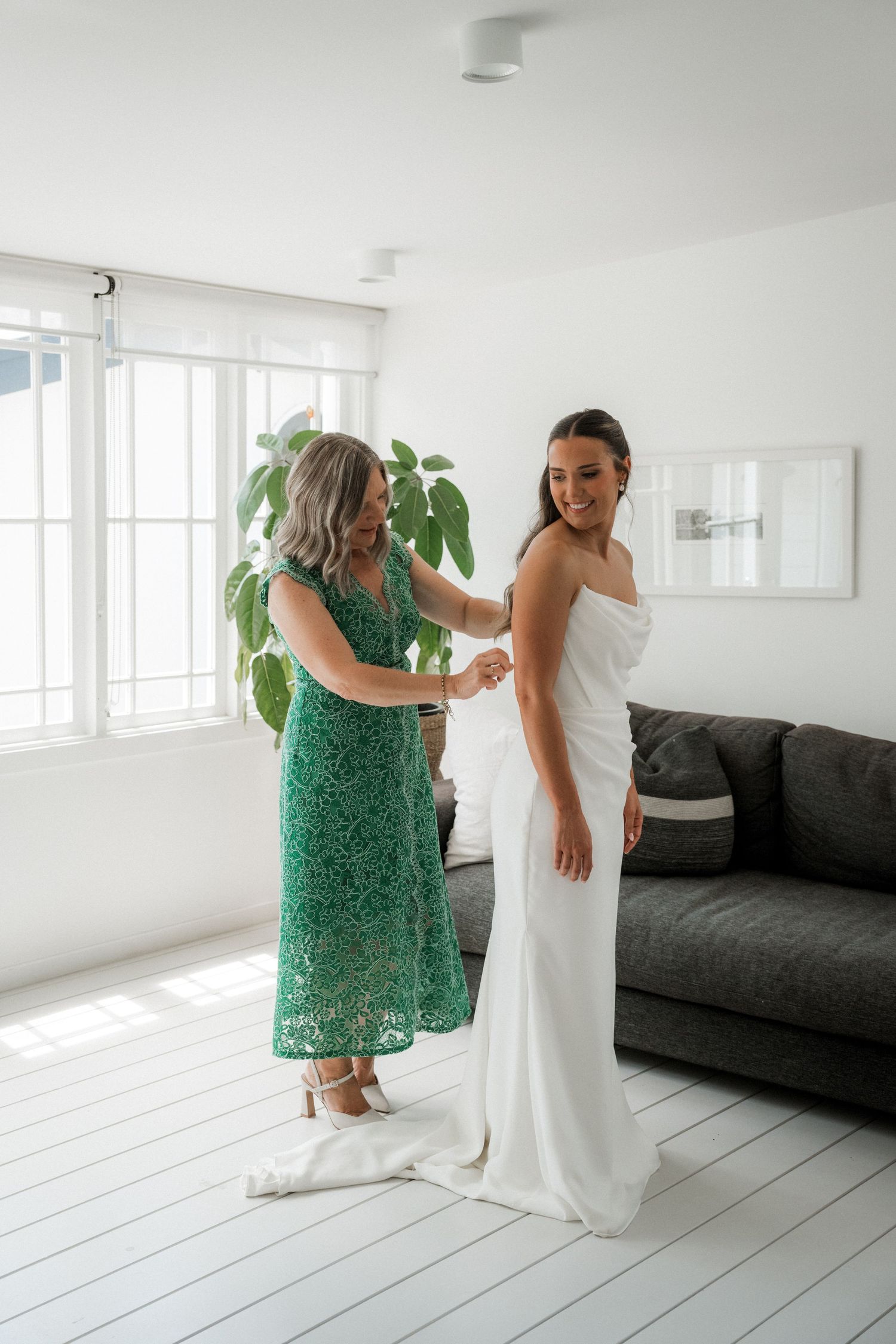 Two women share an emotional moment in a bright white room, one wearing a white gown and another in a green dress.