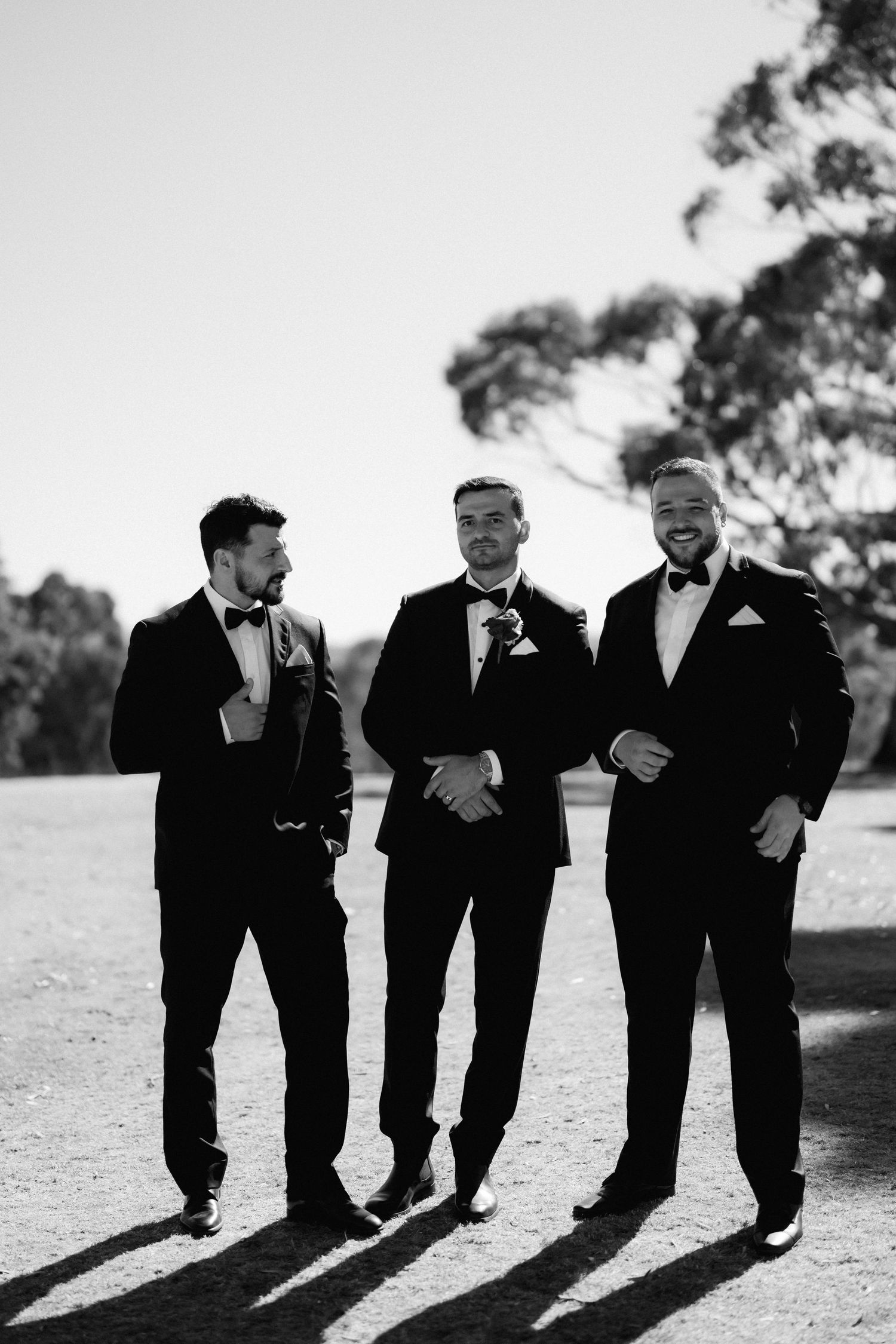 Groomsmen in black tuxedos laughing together while walking outdoors in a candid wedding photo series.