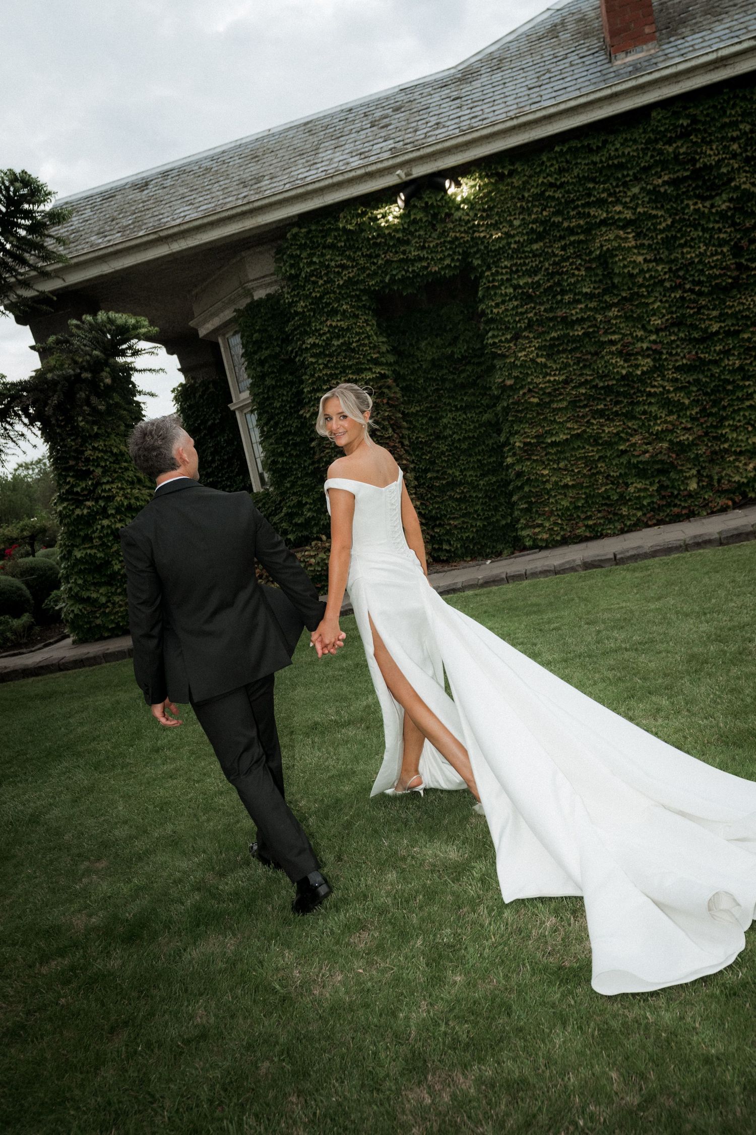 Couple runs playfully across lawn with flowing white dress trailing behind them.