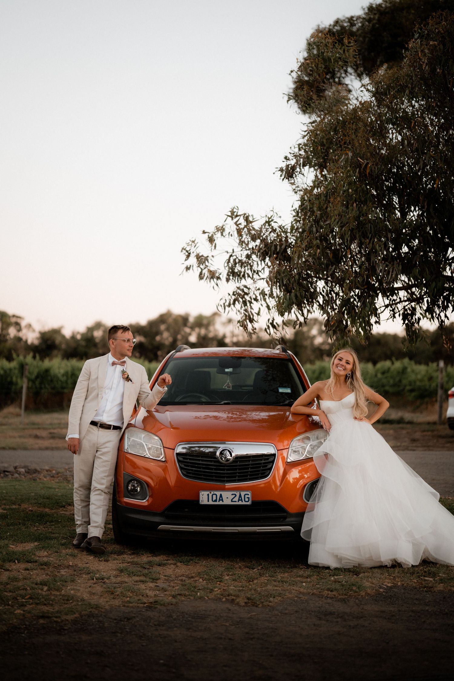 A stylish wedding couple poses with an orange car against a rustic backdrop.