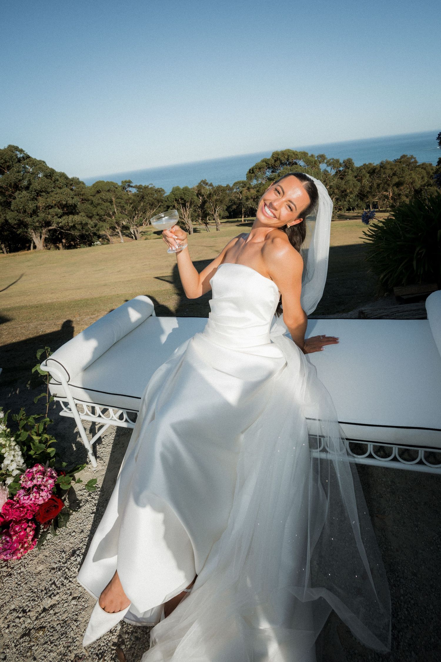 A bride in a flowing white wedding dress poses playfully on a white railing with scenic hills in the background.