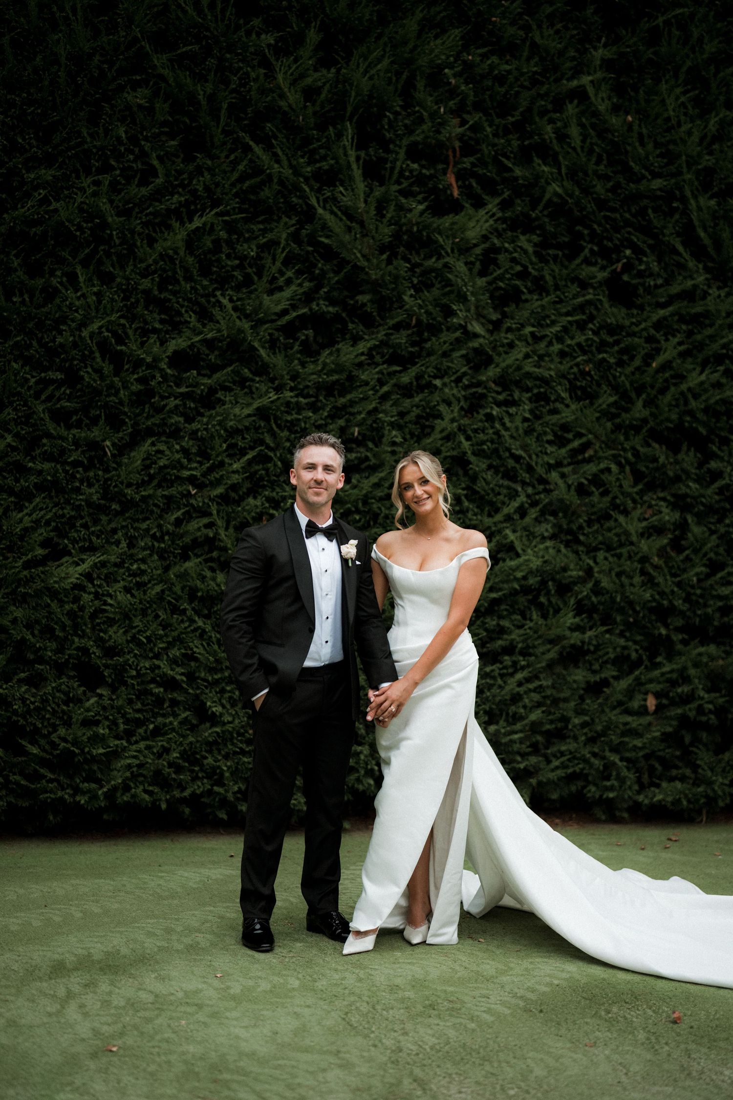 Elegant wedding couple poses against lush hedge backdrop in formal attire.