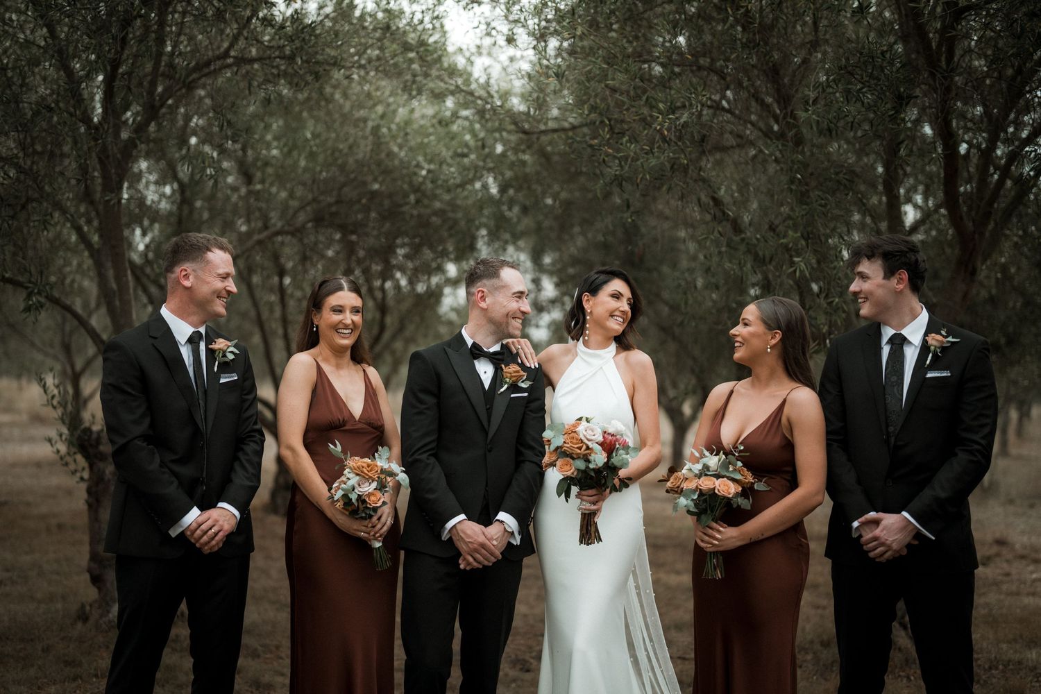 A bridal party in elegant attire stands together outdoors among trees on a moody overcast day.
