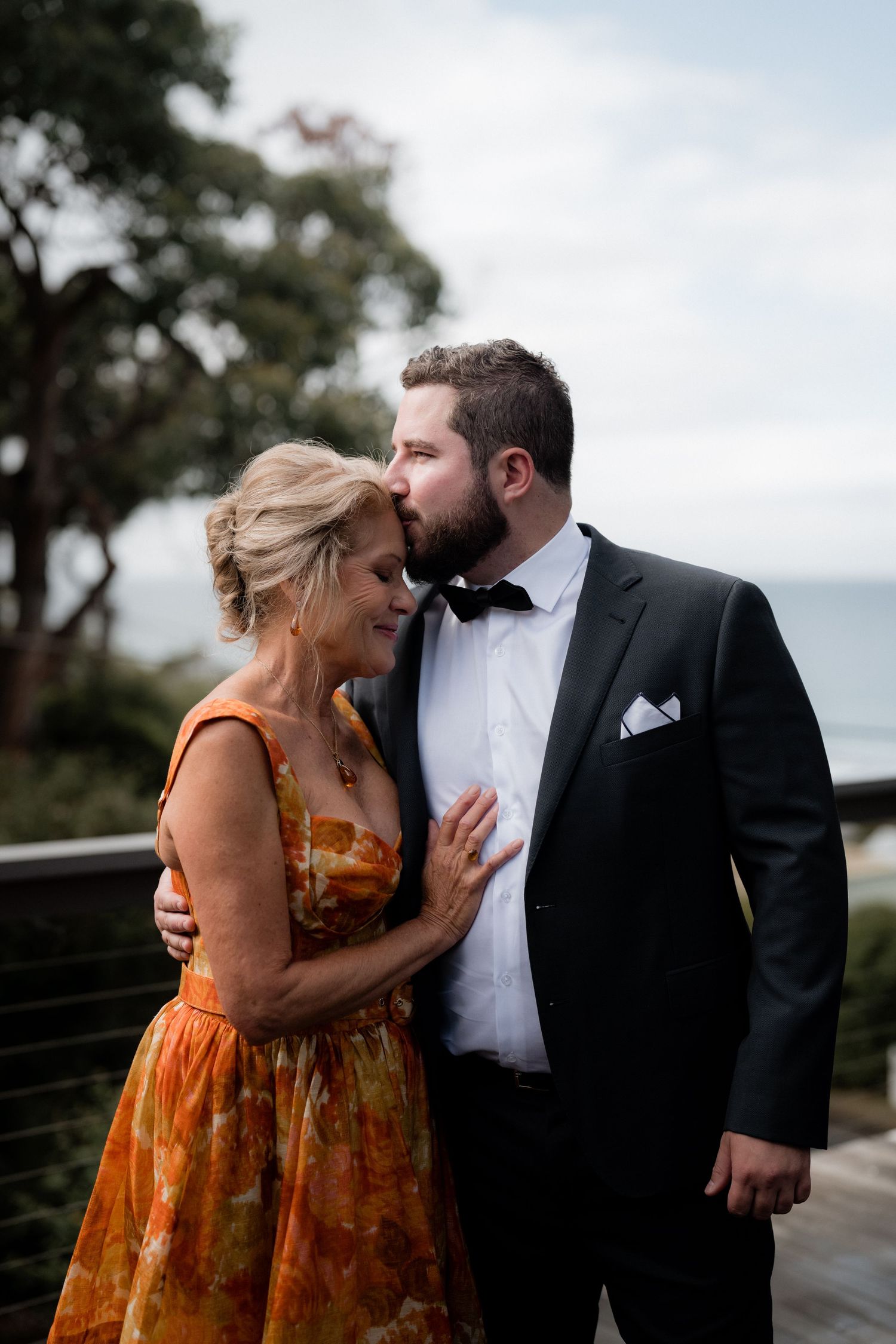 A couple shares an intimate moment outdoors, with her in an orange floral dress and him in a black tuxedo with bow tie.