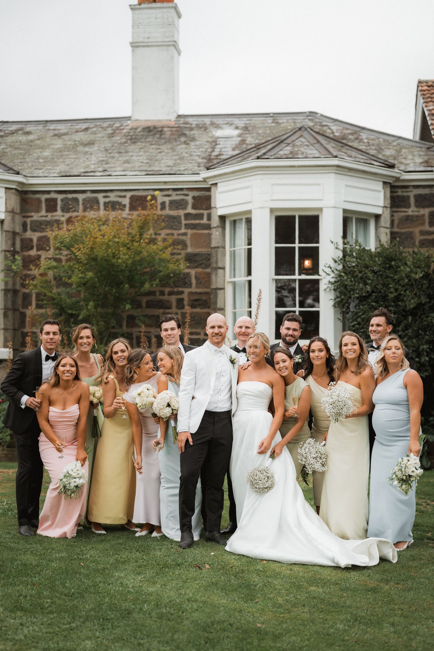 Wedding party poses for formal group photo outside stone building with white trim.