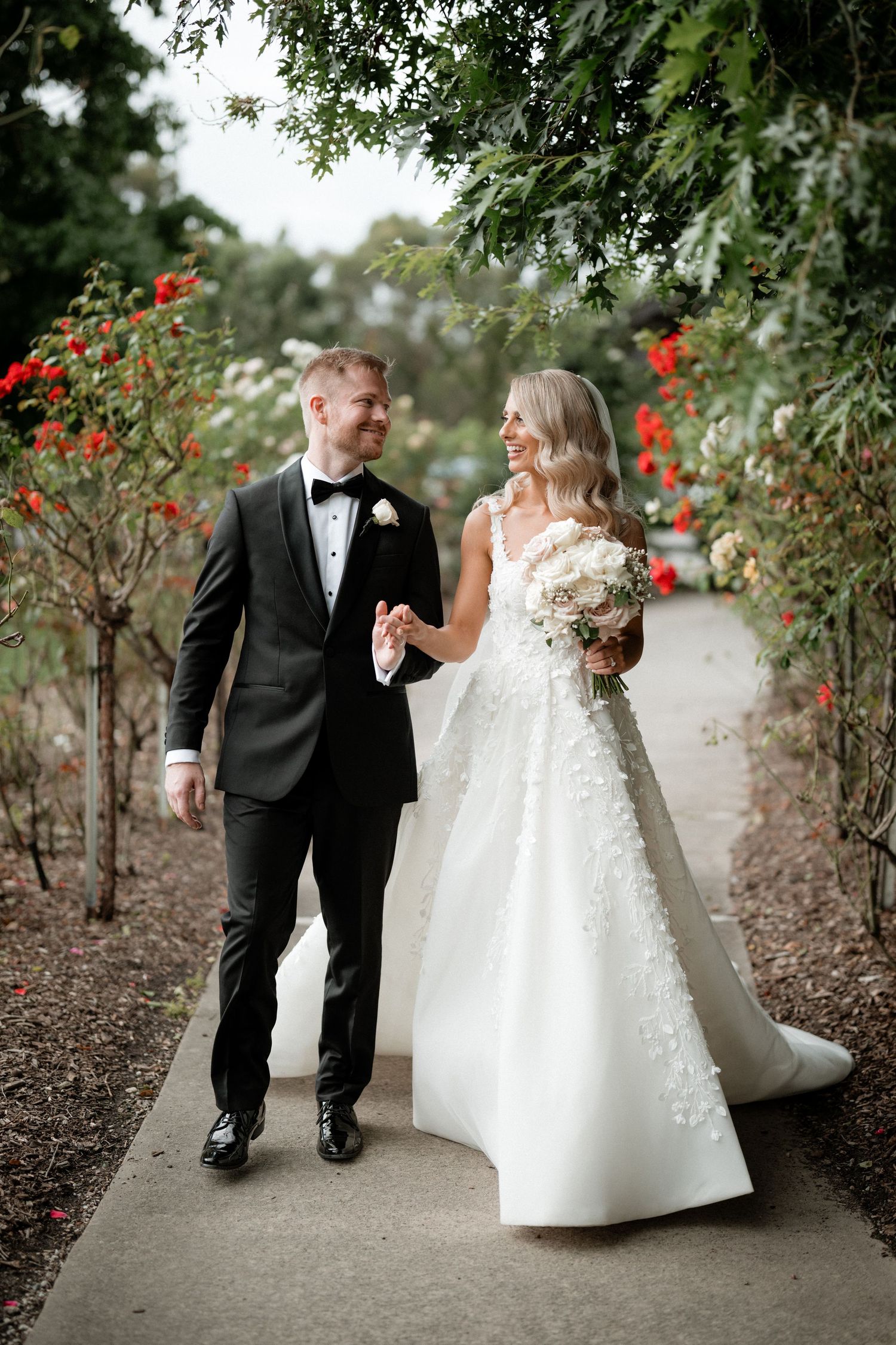 Newlywed couple walking along garden path with red flowers and greenery.