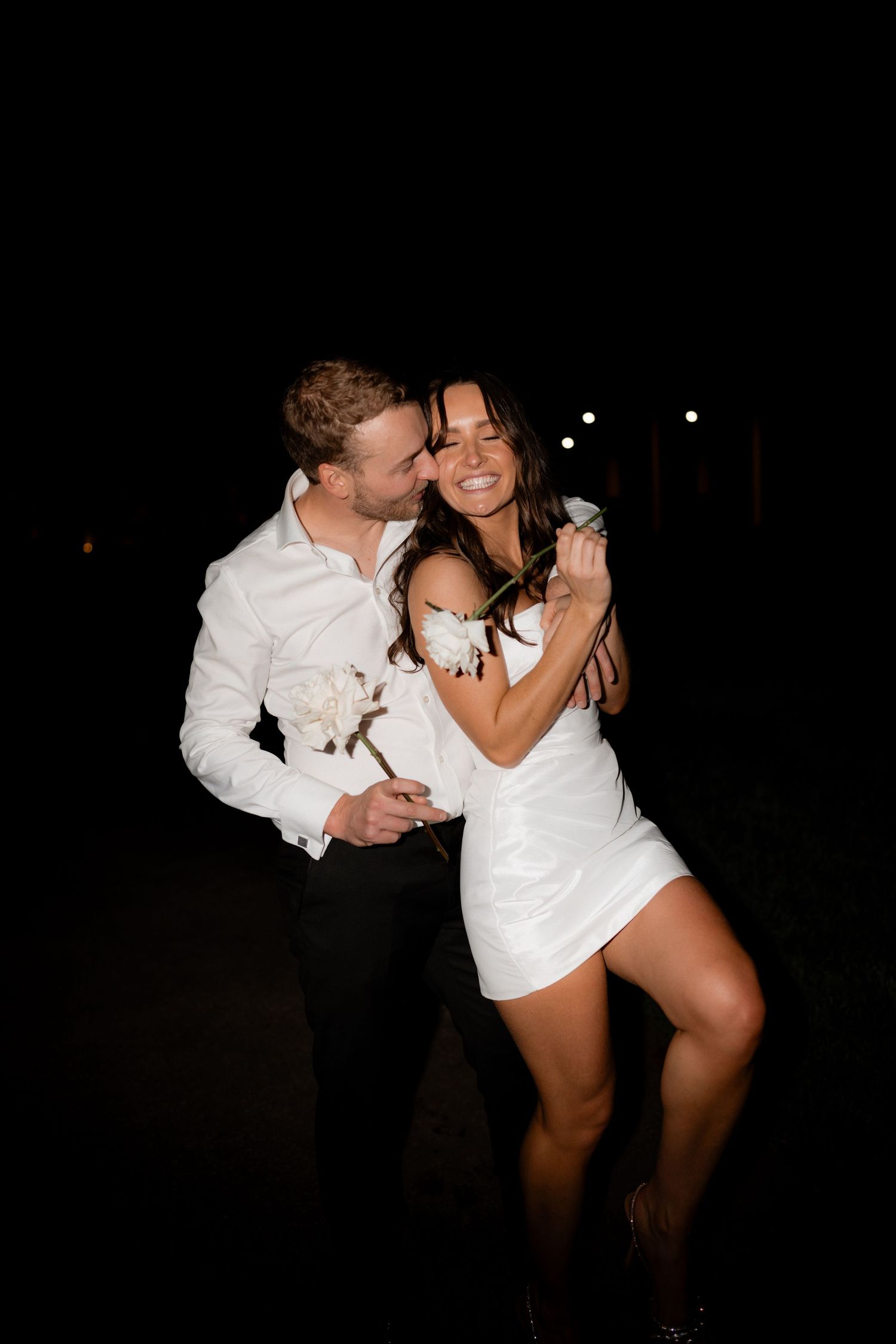 A couple in white outfits shares a romantic nighttime moment together while dancing and embracing.