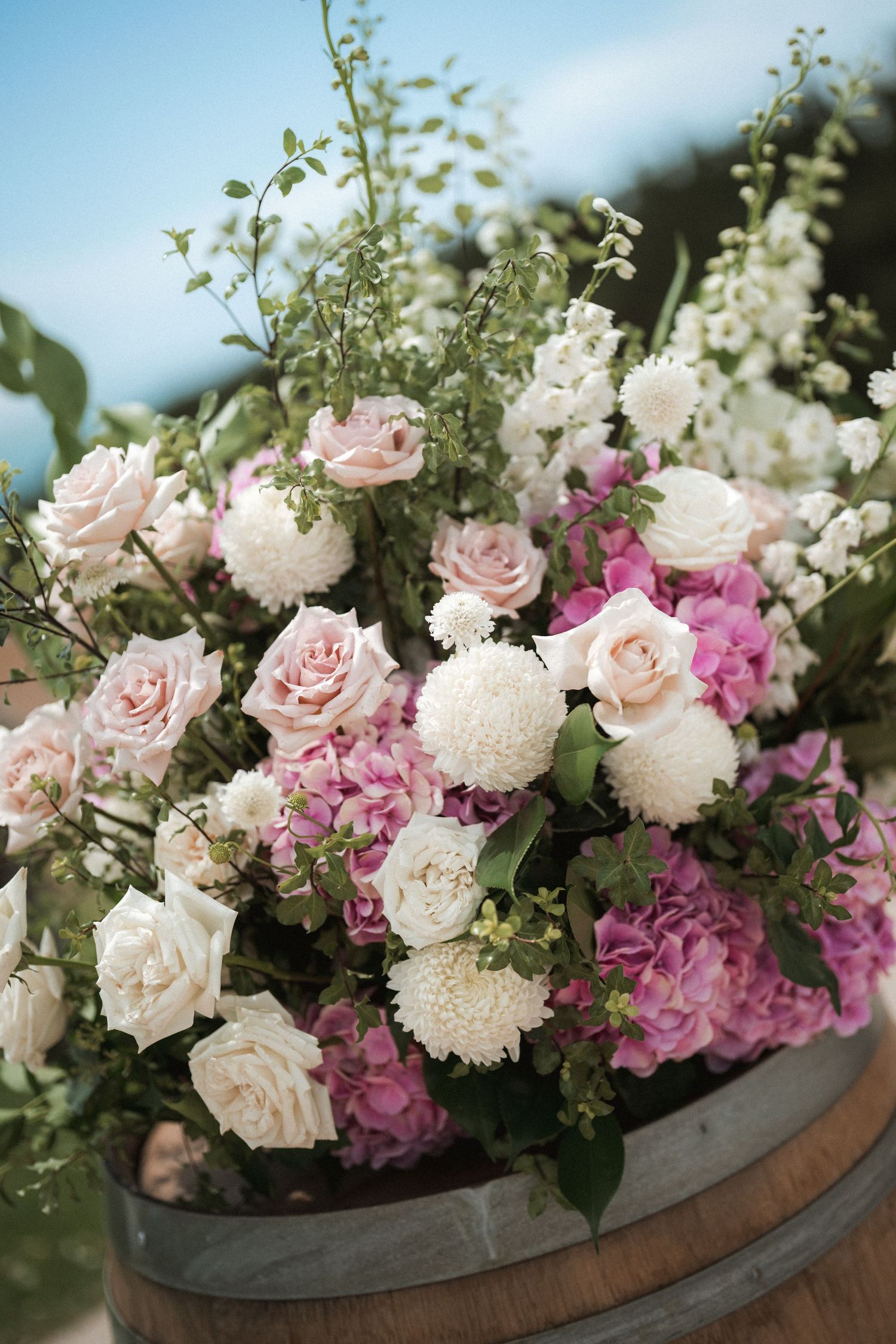 Romantic floral arrangement with pink roses, white dahlias, and hydrangeas displayed in a rustic wooden barrel.