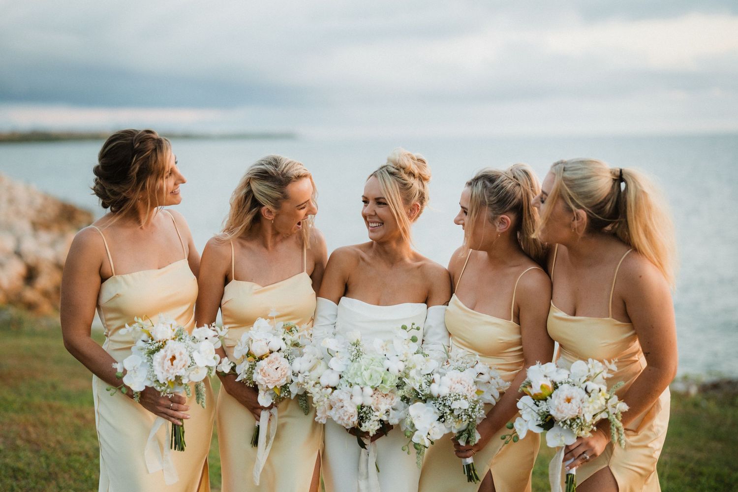 Bridal party in yellow dresses holding white floral bouquets by oceanside cliff.