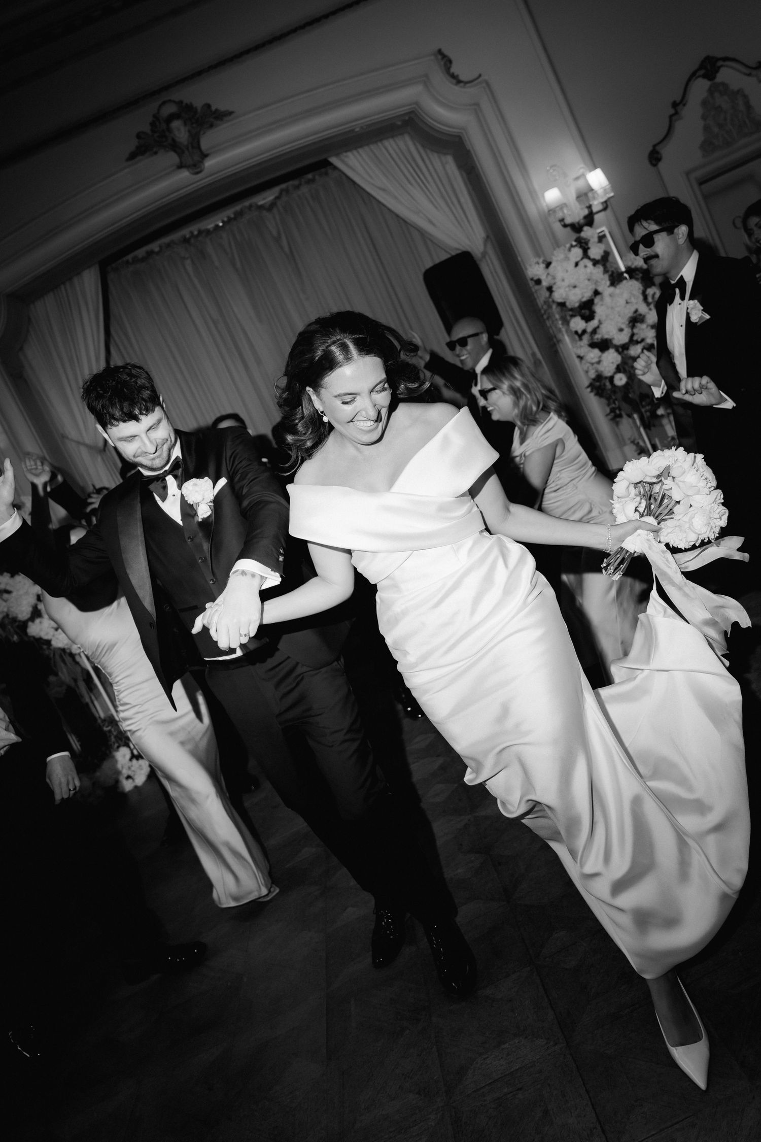 A black and white photo of wedding guests dancing energetically at an elegant indoor reception venue.