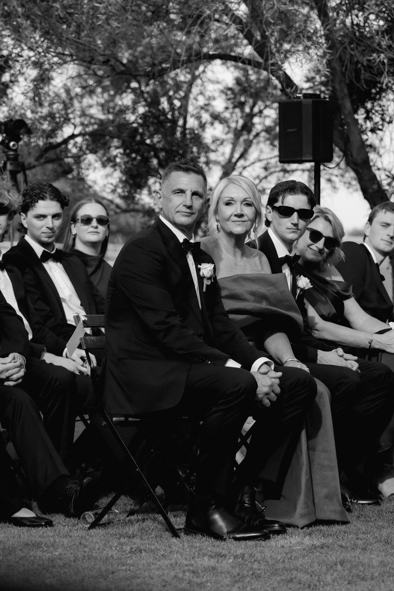 A group of formally dressed guests sitting on grass during an outdoor event in black and white.
