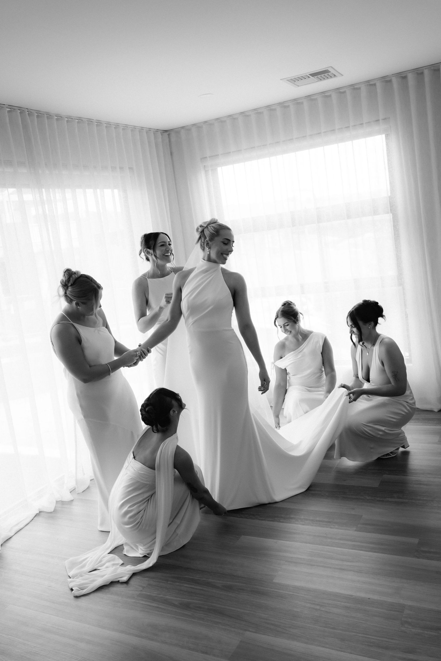 Bridesmaids in white dresses sit on hardwood floor helping with wedding preparations.