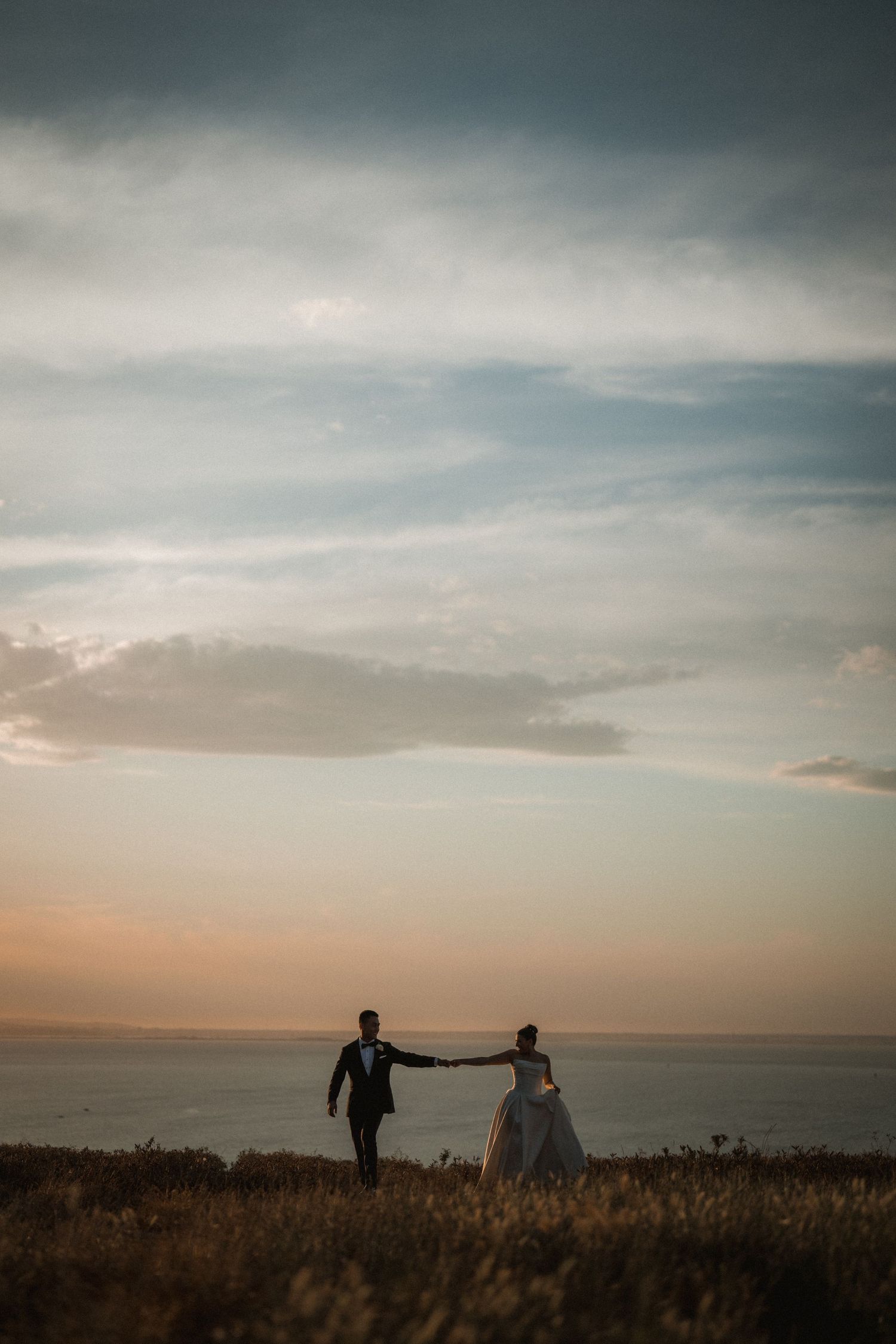 Silhouettes of a couple standing in a field against a dramatic sunset sky over the ocean.