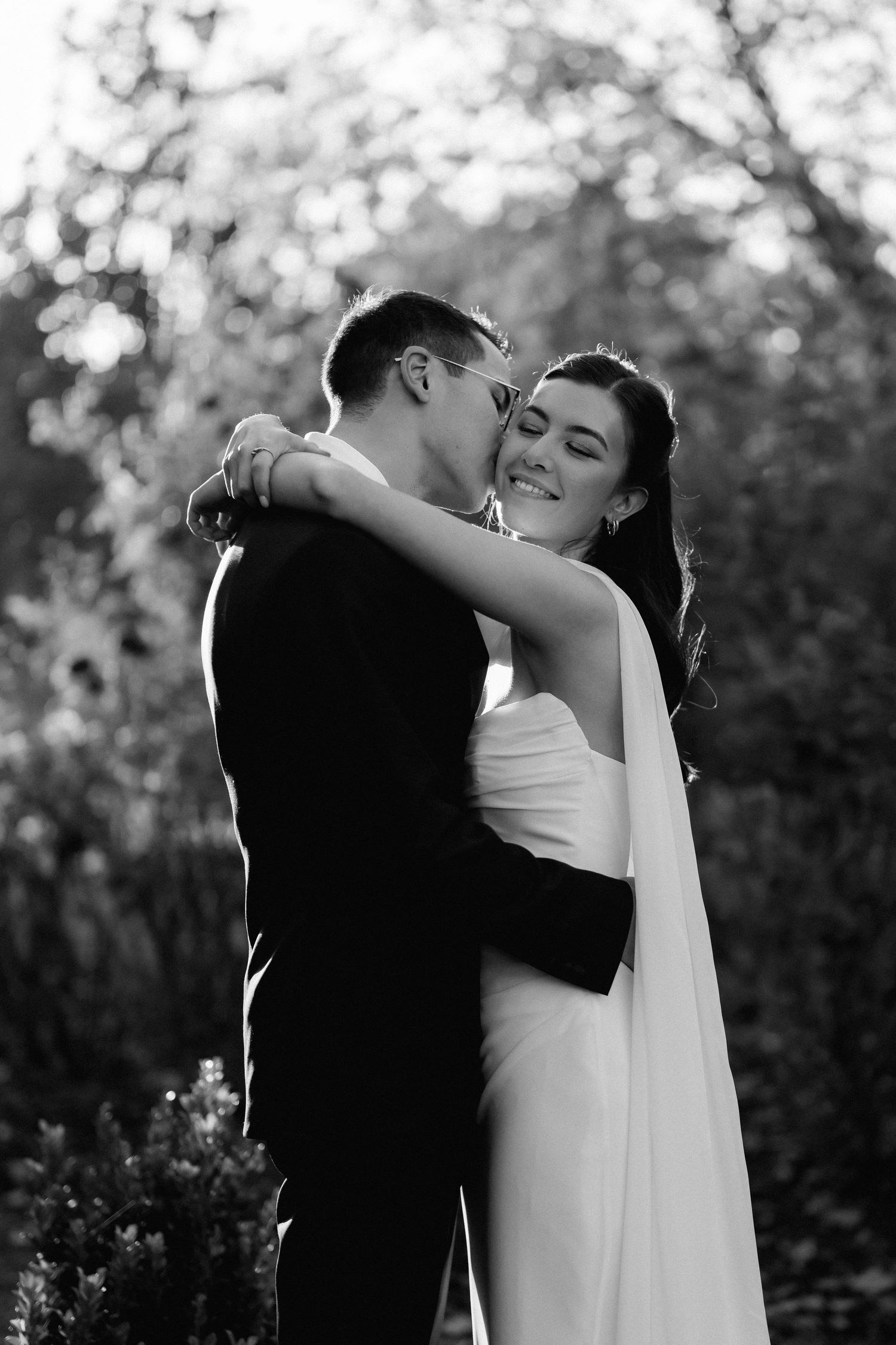 Black and white photo of groom kissing bride on cheek as she smiles in garden setting.