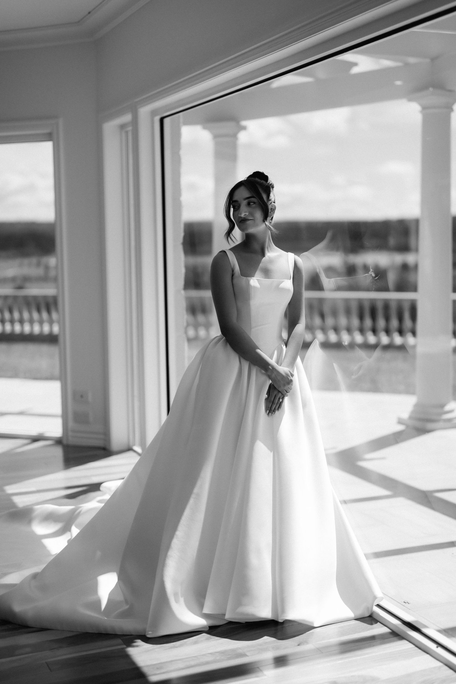 Black and white portrait of a bride in an elegant satin ball gown with a long train standing beside a sunlit window overlooking a terrace.